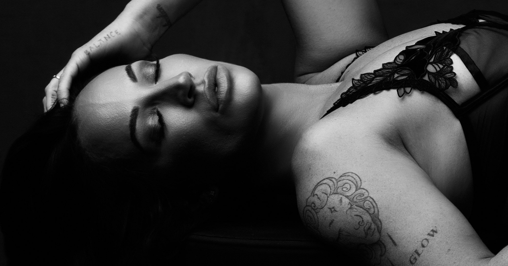 A woman lying down with her eyes closed, holding her head with one hand, wearing a black lace top, with tattoos visible on her arm and hand, in black and white photography.
