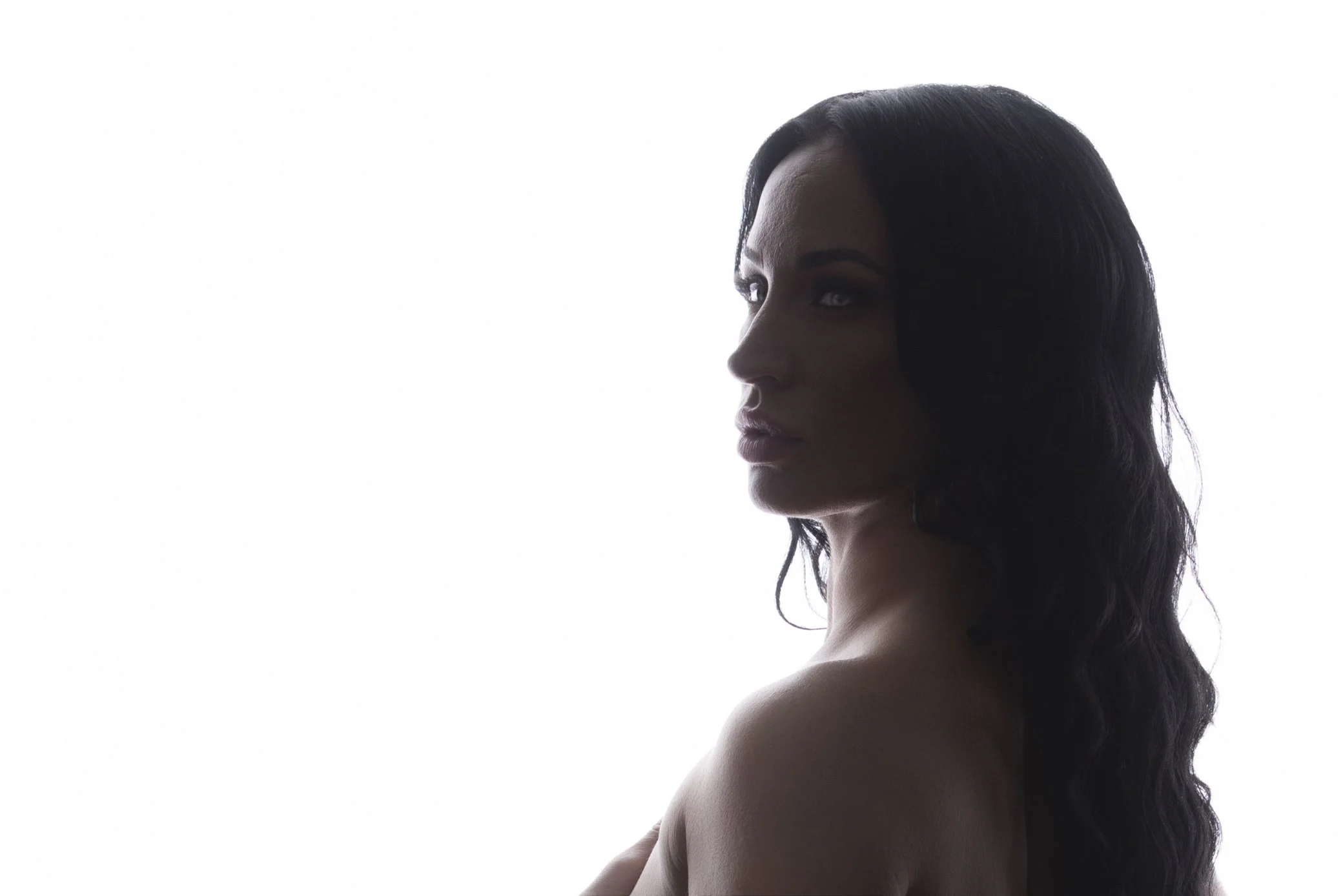 A side profile of a woman with long dark wavy hair, facing left, with minimal background.