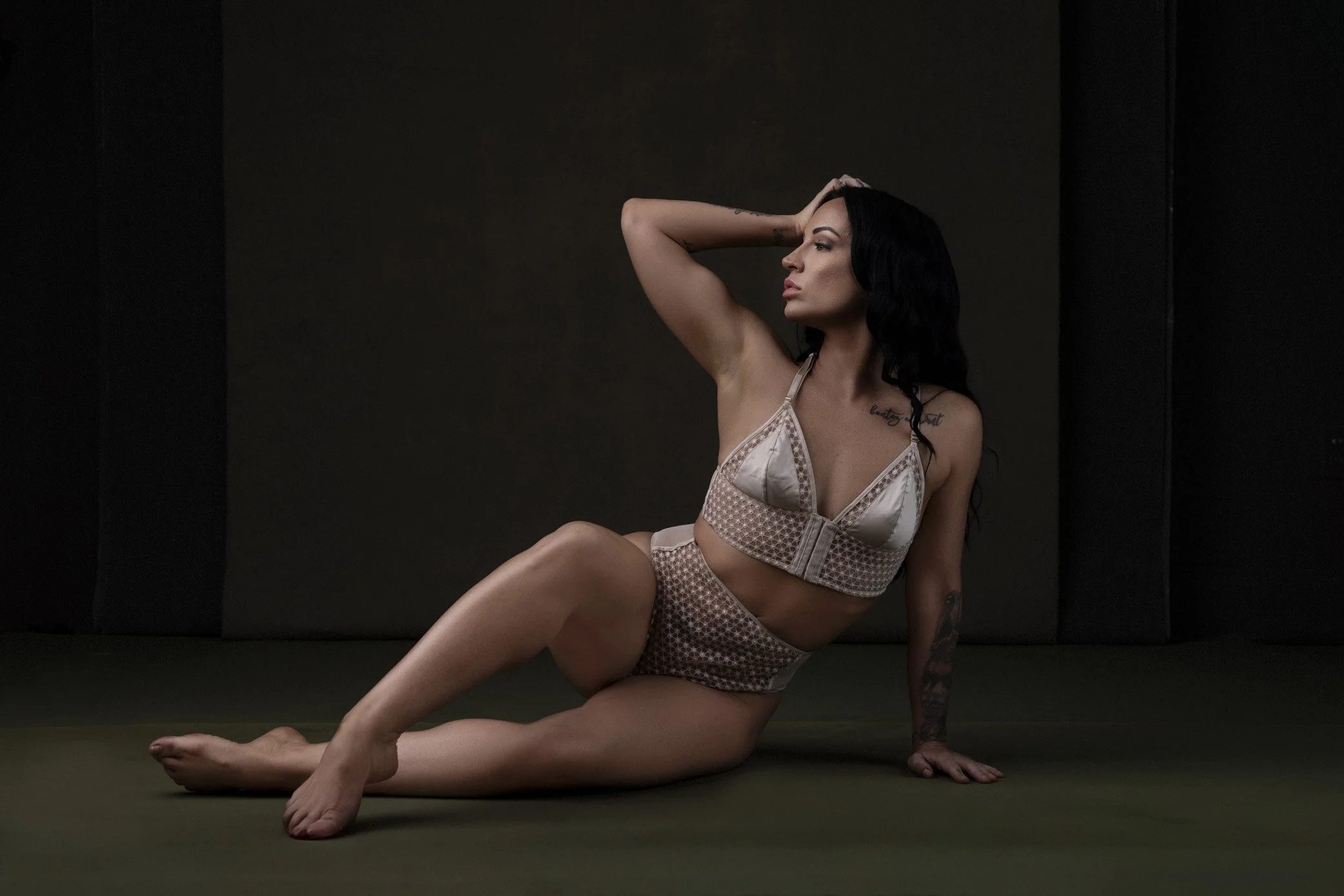 A woman with dark hair and tattoos poses on a dark floor, wearing beige lingerie, sitting with one arm behind her head and the other hand on the ground.