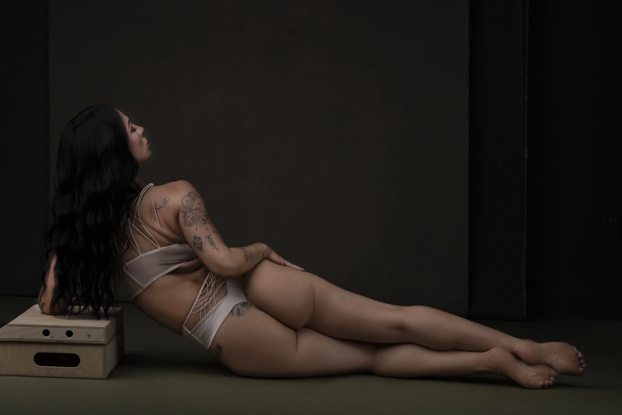 A woman with long black hair and tattoos, wearing revealing beige lingerie, reclining on a mat with her head resting on a small wooden box, against a plain dark background.