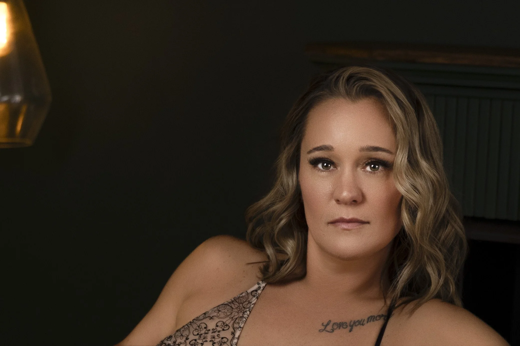 A woman with wavy brown hair and makeup, wearing a lacy top, sitting indoors with a dark background
