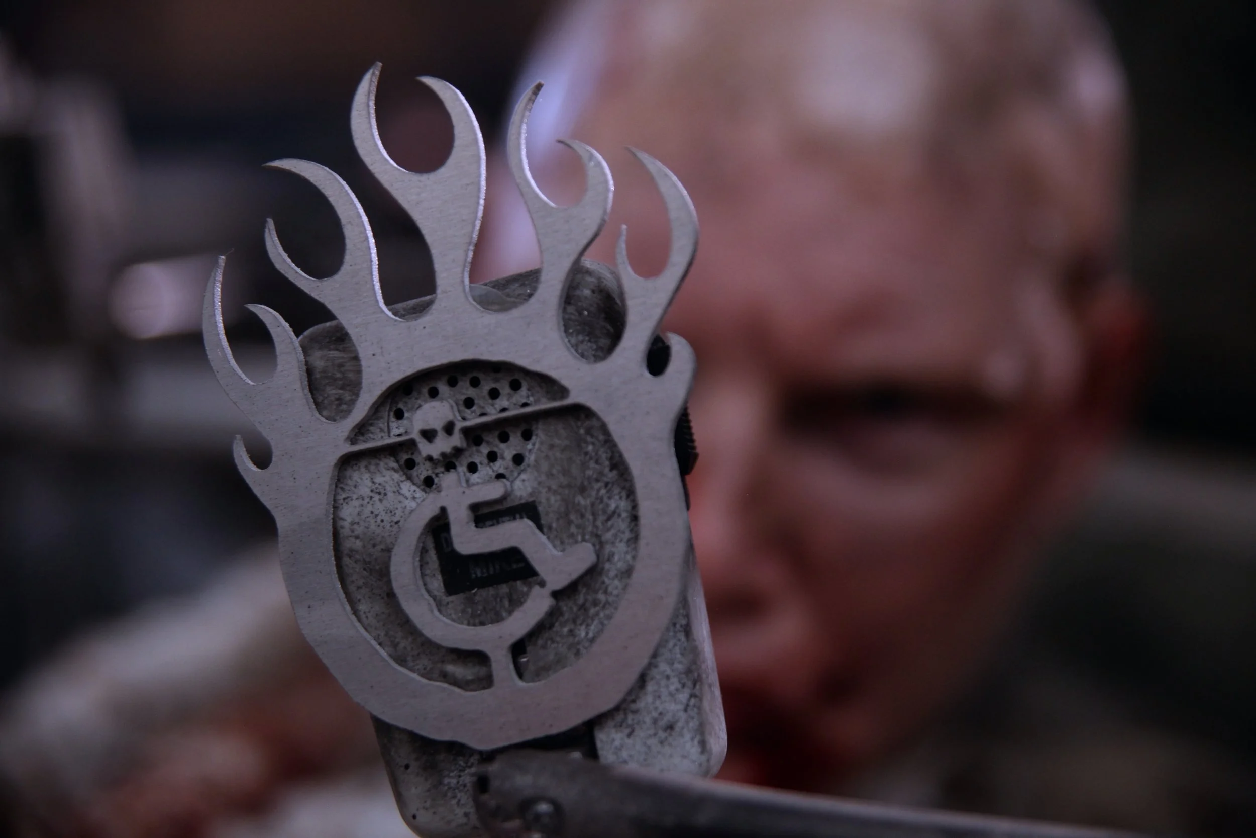 My metal flaming wheelchair symbol is welded to a microphone, which is near my face. My filthy face is blurred in the background.