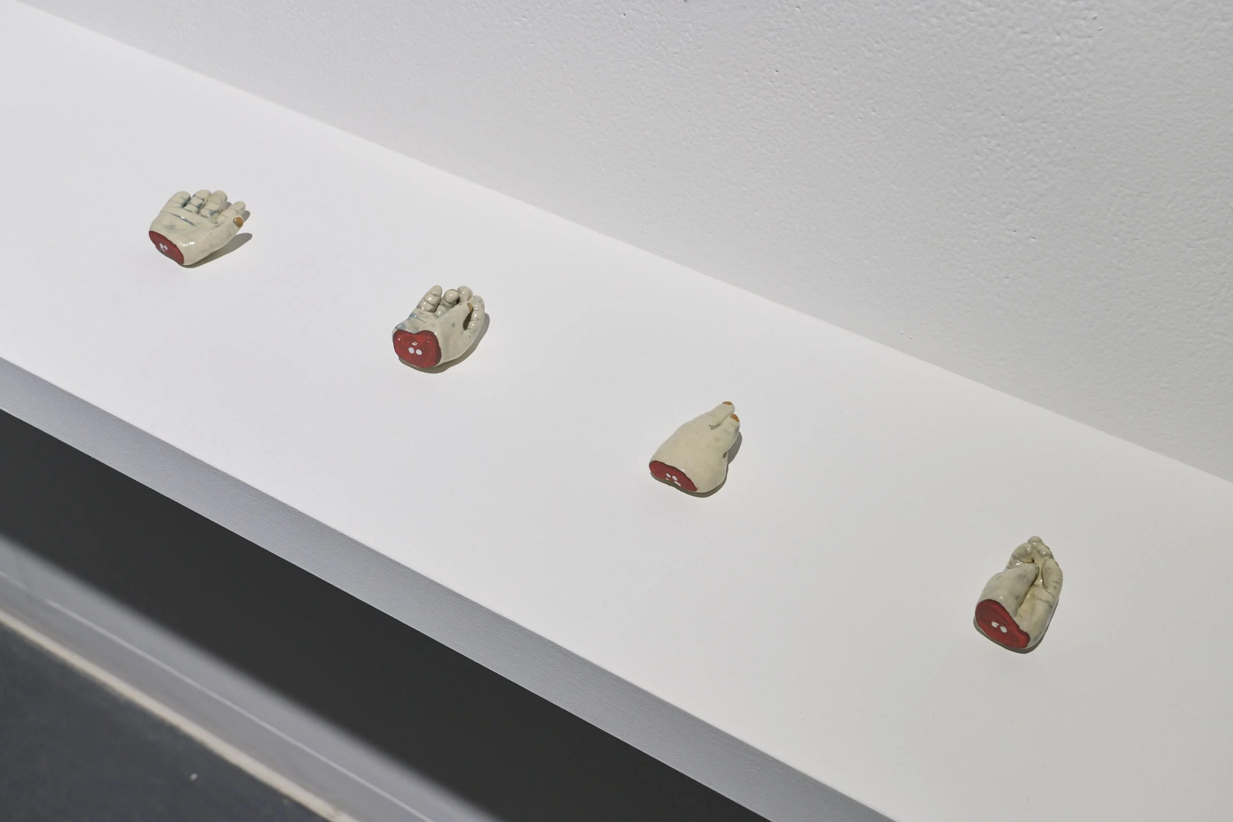 A white shelf with 4 ceramic objects which reference severed infant hands, the bones can be seen in the severing at the wrist. The hands have pale skin and are in various states of gesturing