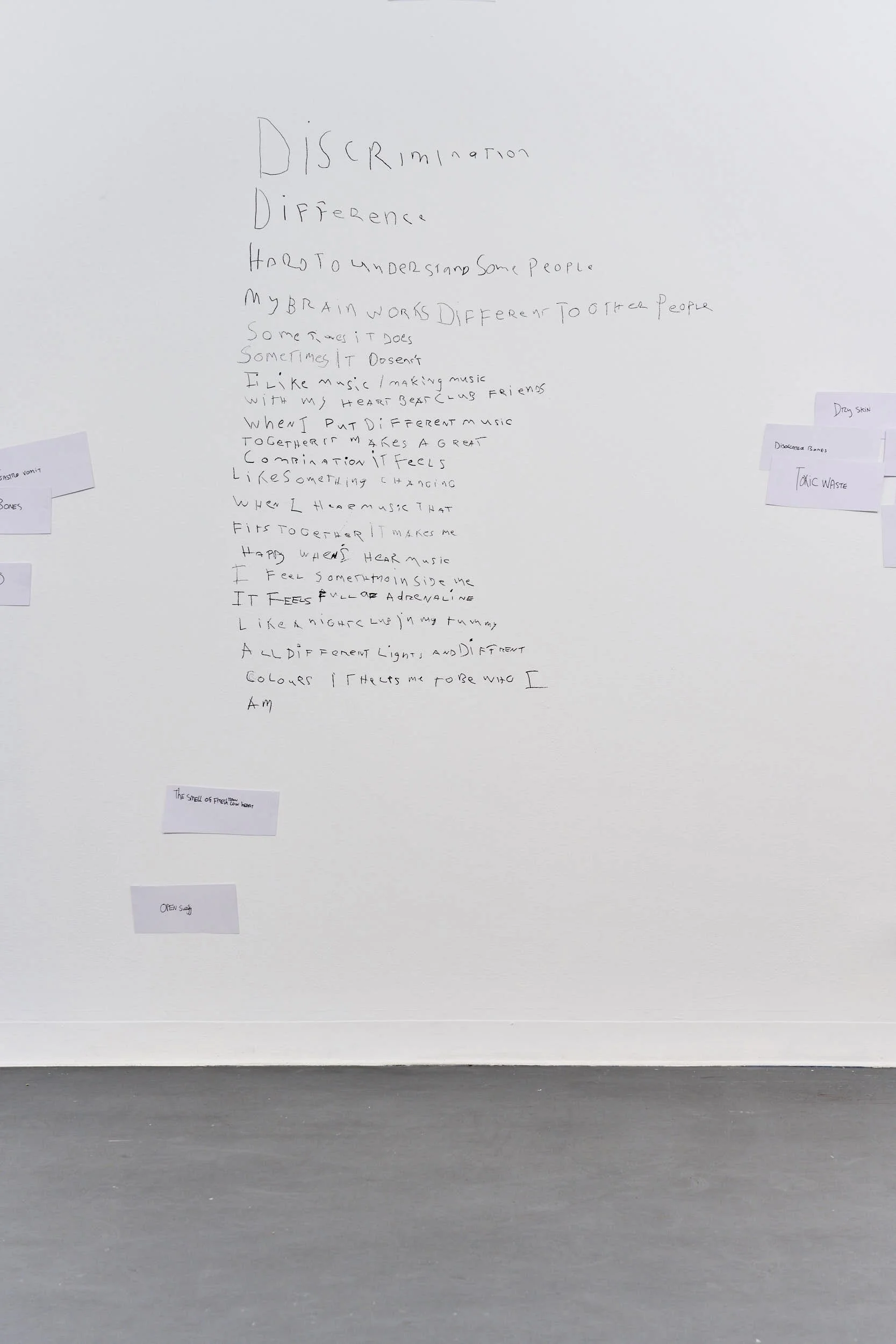 Wide view of the poem with scattered pieces of rectangular paper layered on top, stuck on the wall, surrounding the poem, reading, “DRY SKIN,” “TOXIC WASTE,” “DISLOCATED BONES,” “OPEN surgery”.