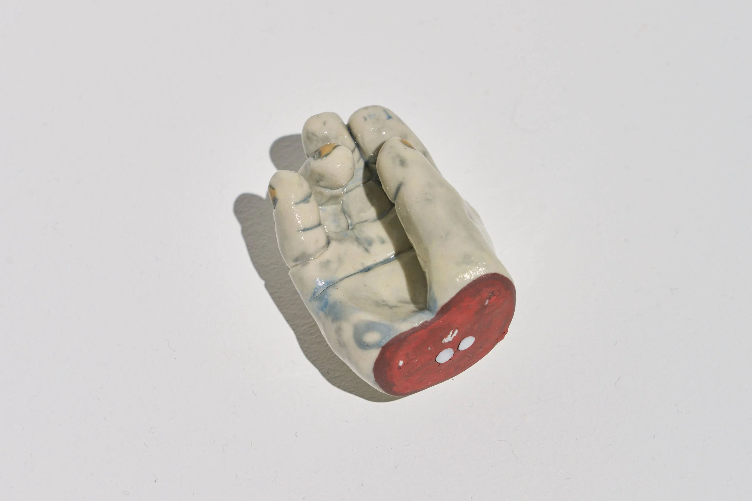 Close up view of the ceramic object reveals a glazed complexion with blue tones in the wrinkles, yellow nails, and a red cross section.  The hand is gesturing with the palm up with the thumb finger lightly touching the index finger.