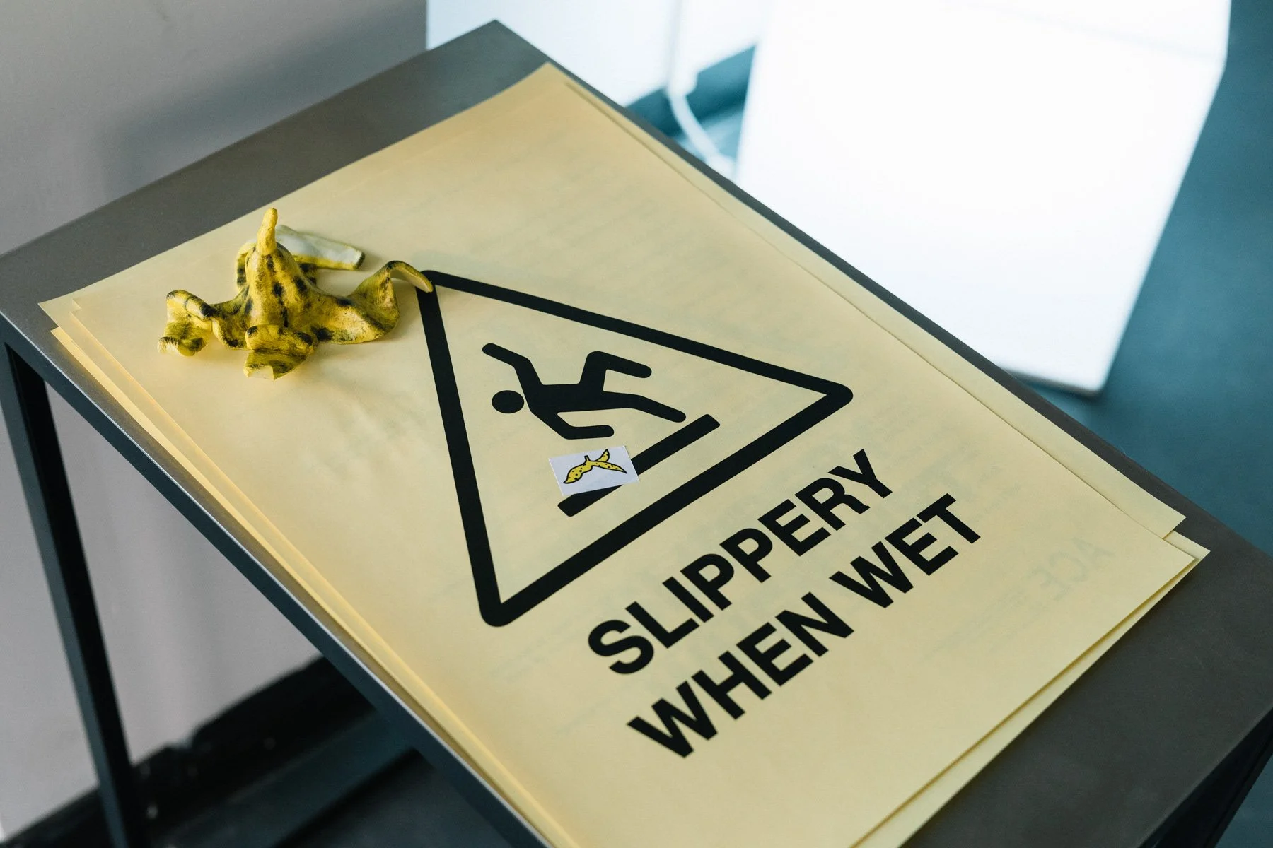 A pile of yellow posters reading “slippery when wet” with a graphic of a person slipping on a little banana peel sticker that has been added. The posters are held down by a banana skin ceramic sculpture.