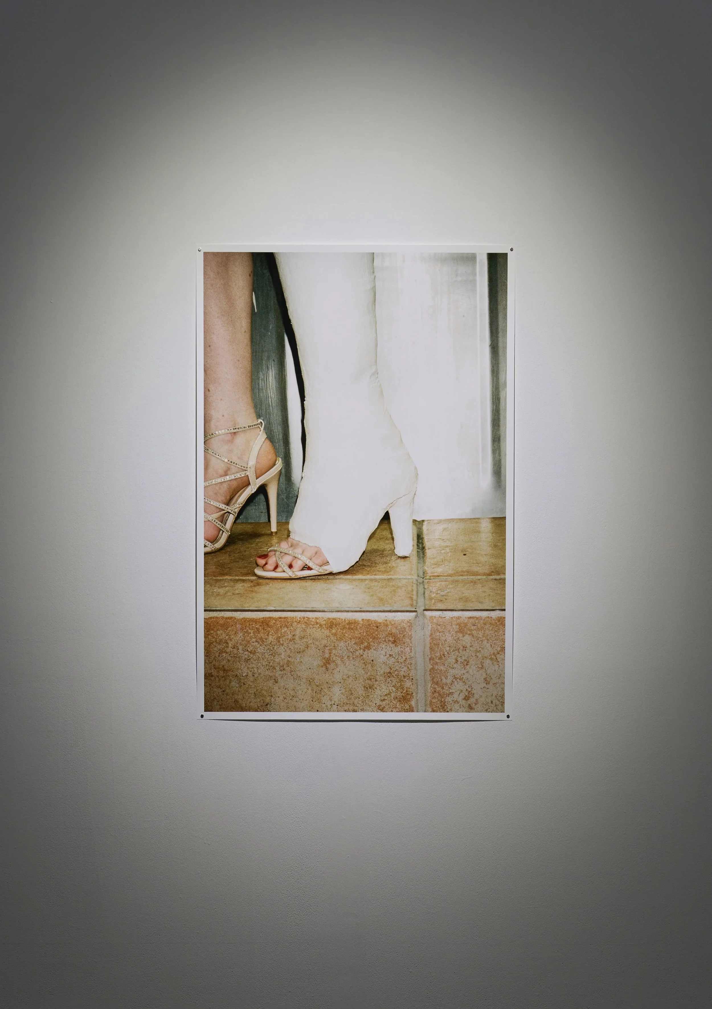 A photo depicting a pair of legs standing on the tiles of a urinal floor. The right leg is in a plaster cast, also encasing a gold stiletto on the foot. The toes are exposed and red nail polish shines out