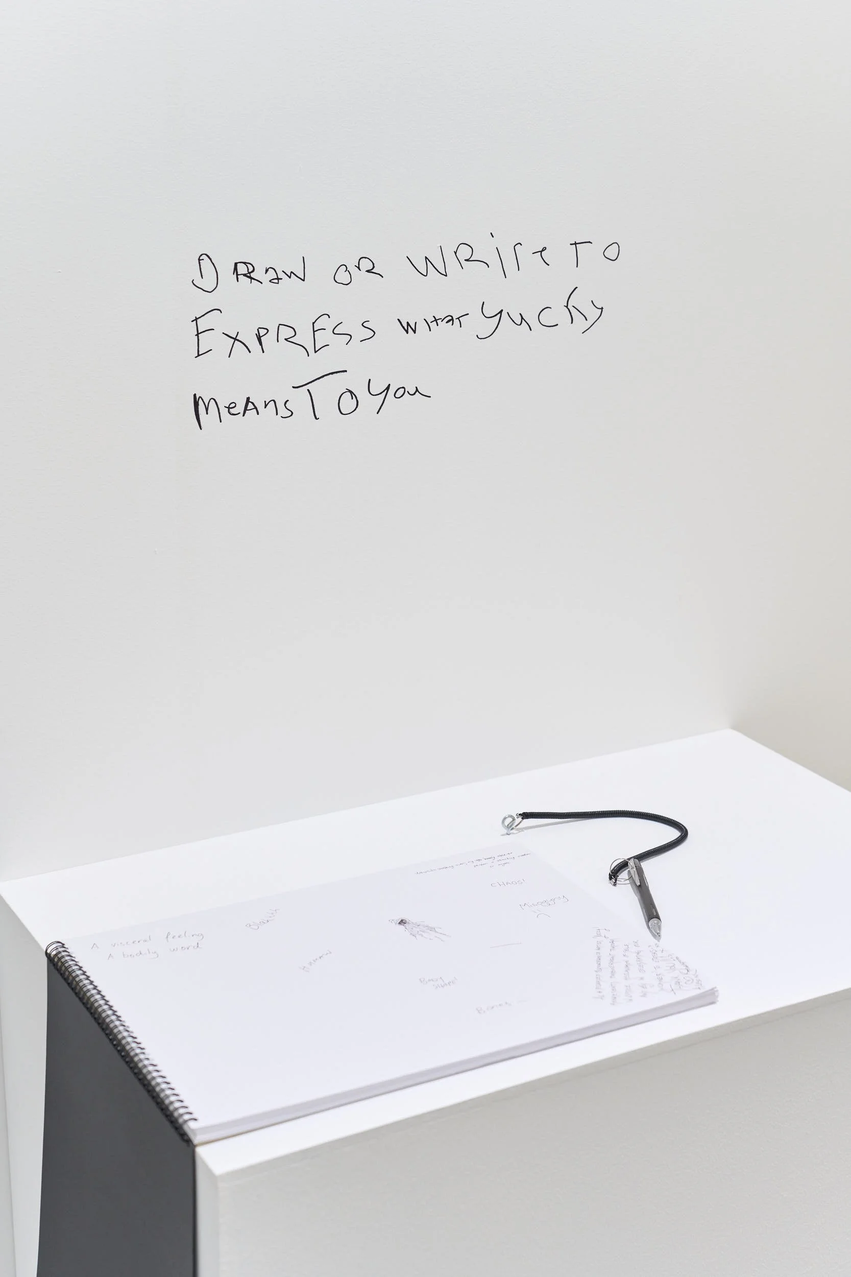 Written on a white wall with black pencil reads, “DRAW OR WRITE TO EXPRESS WHAT YUCKY MEANS TO YOU,” above a white table with an open notepad and a pen attached to it.