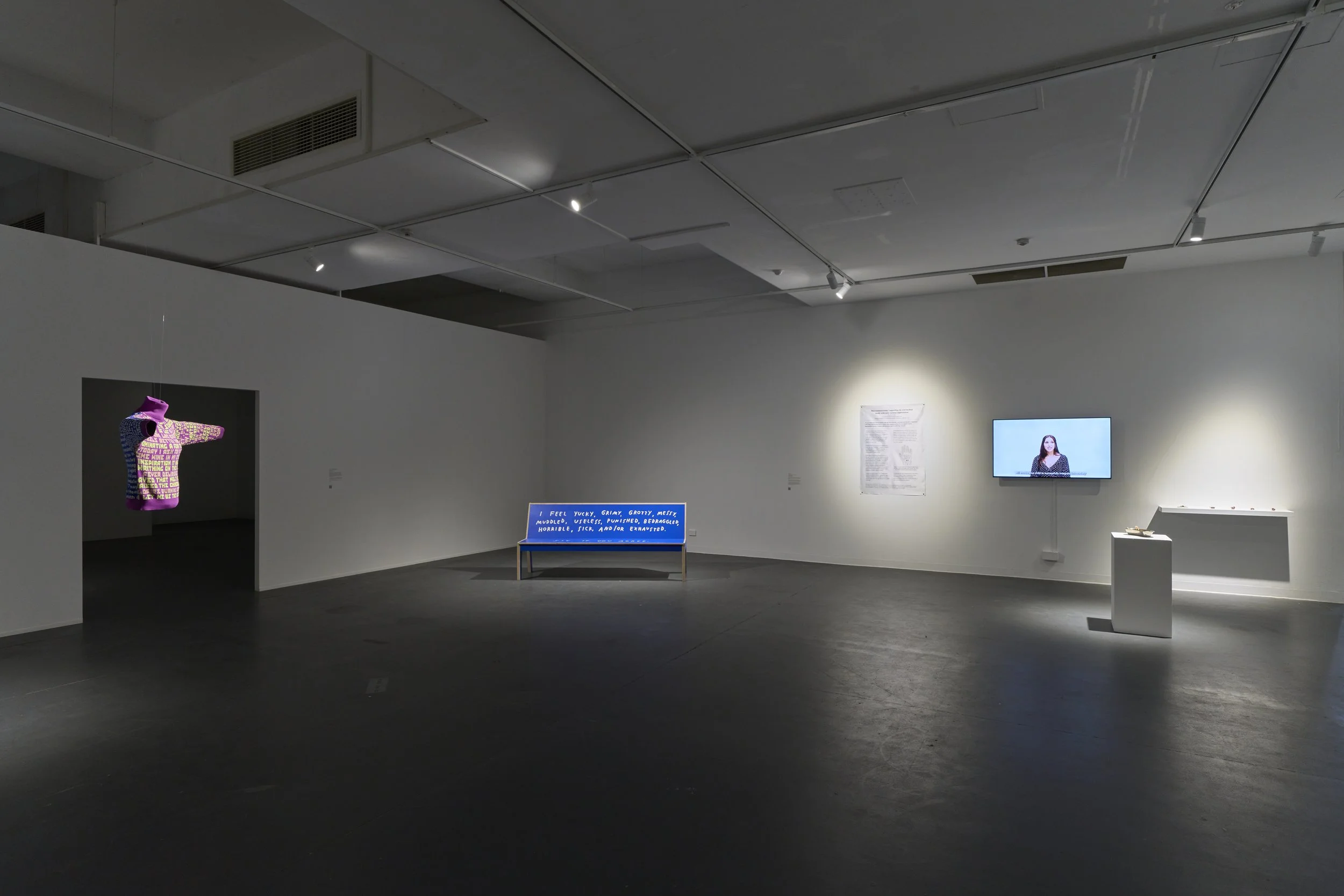 Back in the first room there is Makeda Duong Jumper. In the middle there is a blue bench To the right, there is white fabric on the wall. next to is a tv screen. There is a plinth and shelf to the right of the tv screen.