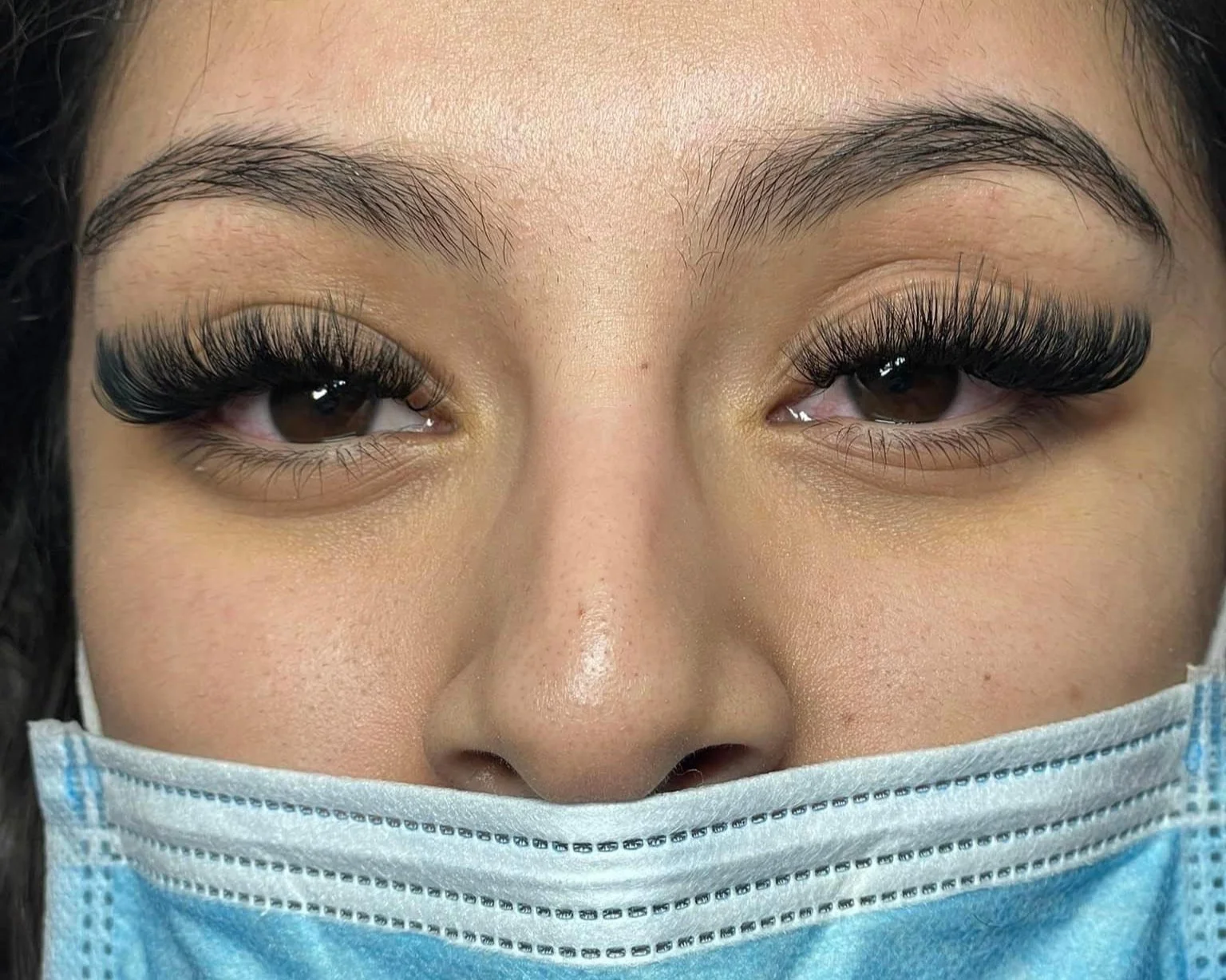 Close-up of a woman's face with brown eyes, wearing dramatic false eyelashes and a blue disposable face mask.