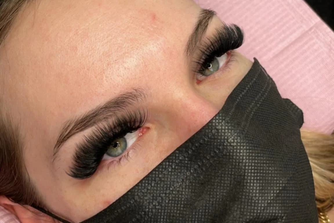 Close-up of a woman lying down, wearing a black face mask, with long, thick eyelash extensions and natural eyebrows.