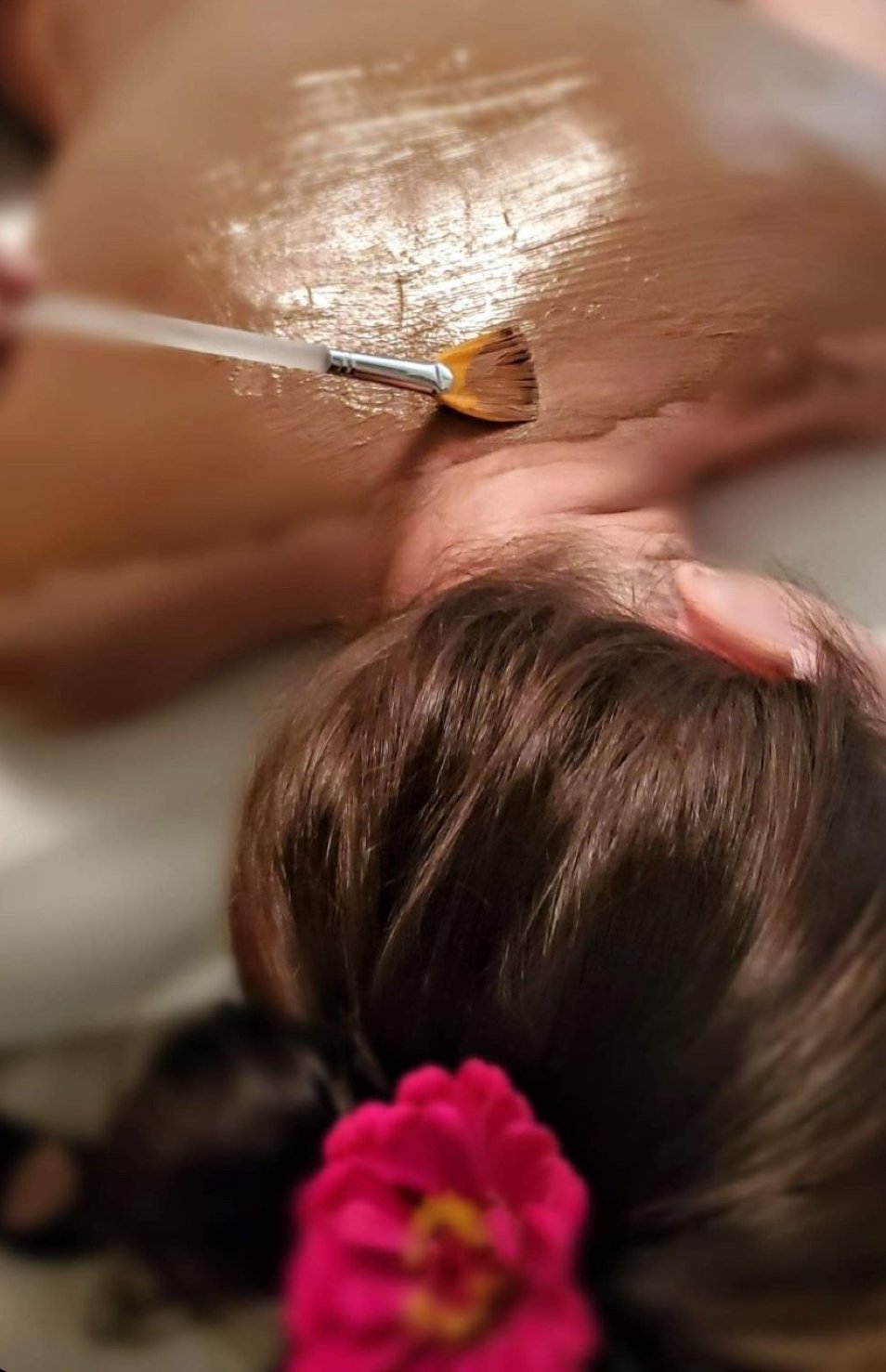 Person receiving a facial or skincare treatment with a brush applicator on their face, lying down with brown hair and a pink flower in their hair.