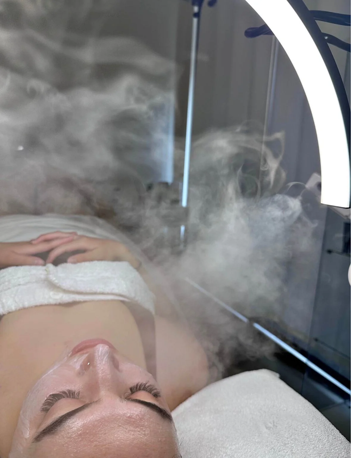 A woman lying down inside a steam facial cabinet with steam fog surrounding her, her eyes closed and face relaxed.