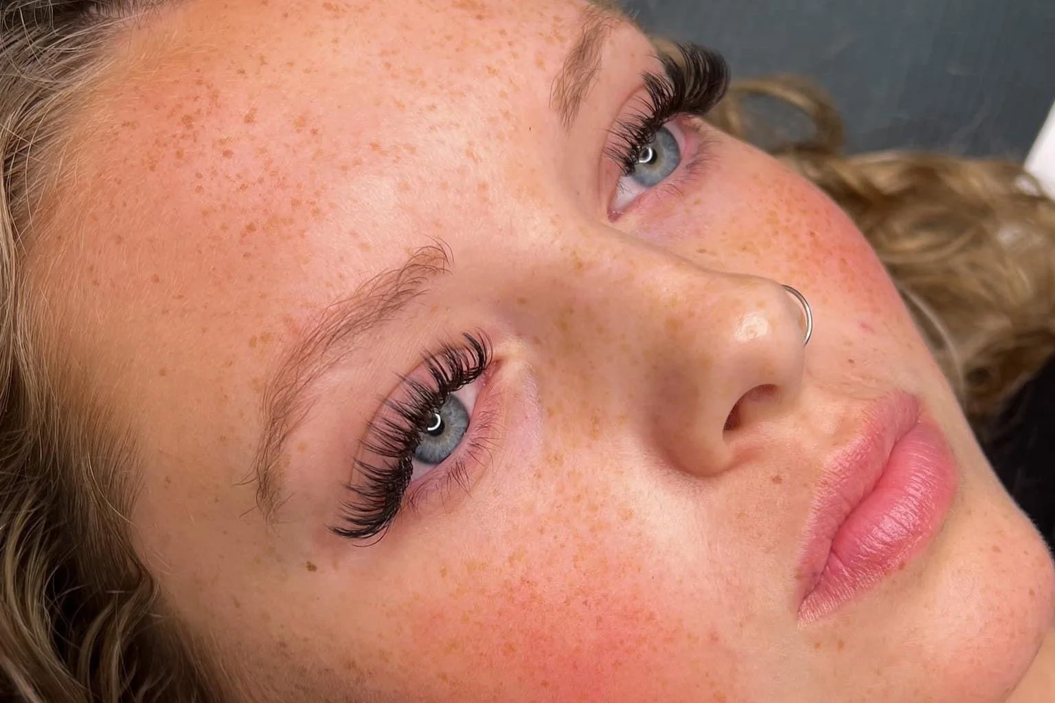 Close-up of a young woman with blue eyes and light freckles, lying down with her head turned slightly to the side, showing her eyelashes, nose, and lips.