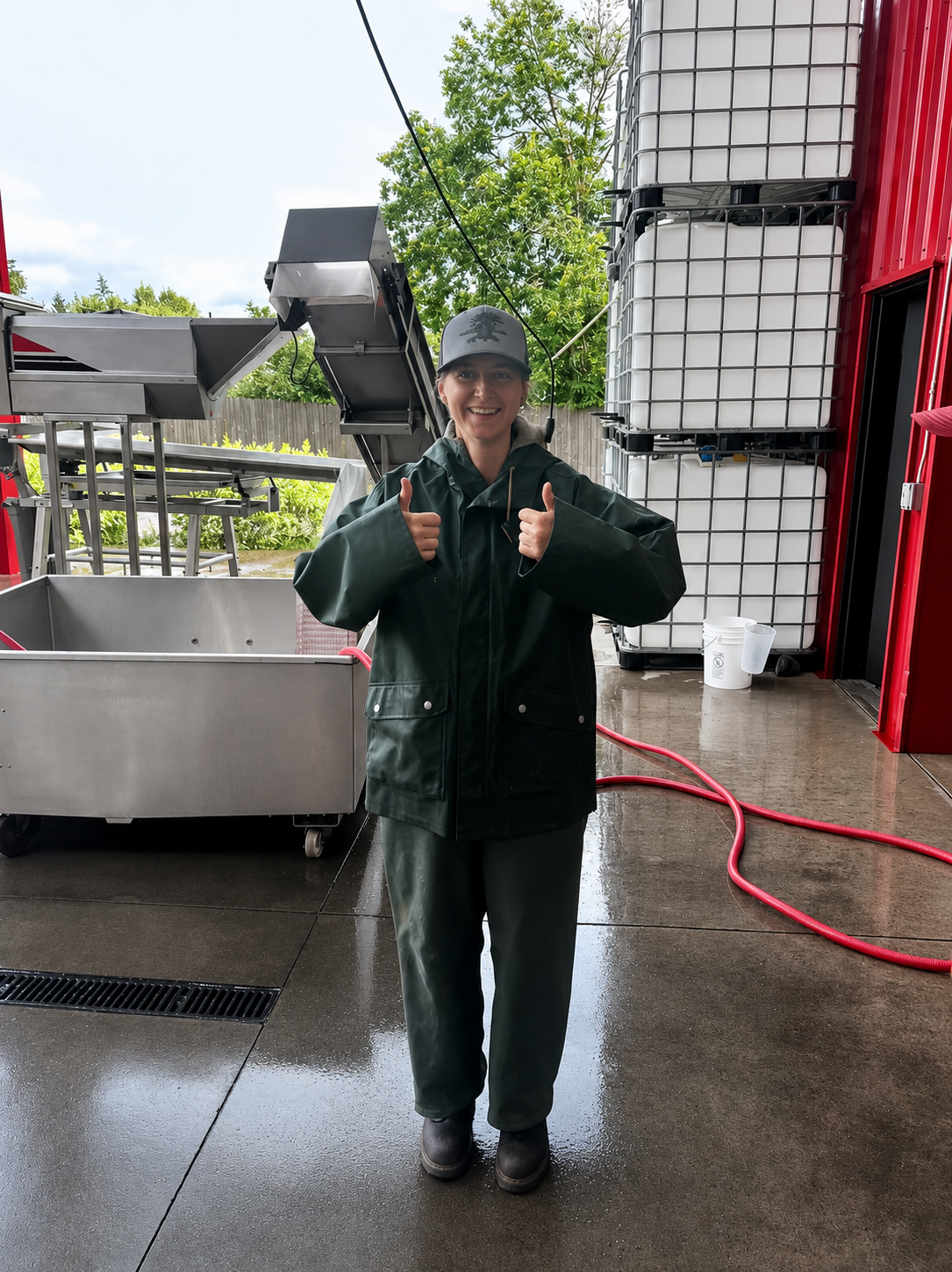 Woman smiling and giving thumbs up in a wet, industrial setting with large plastic tanks and equipment behind her.