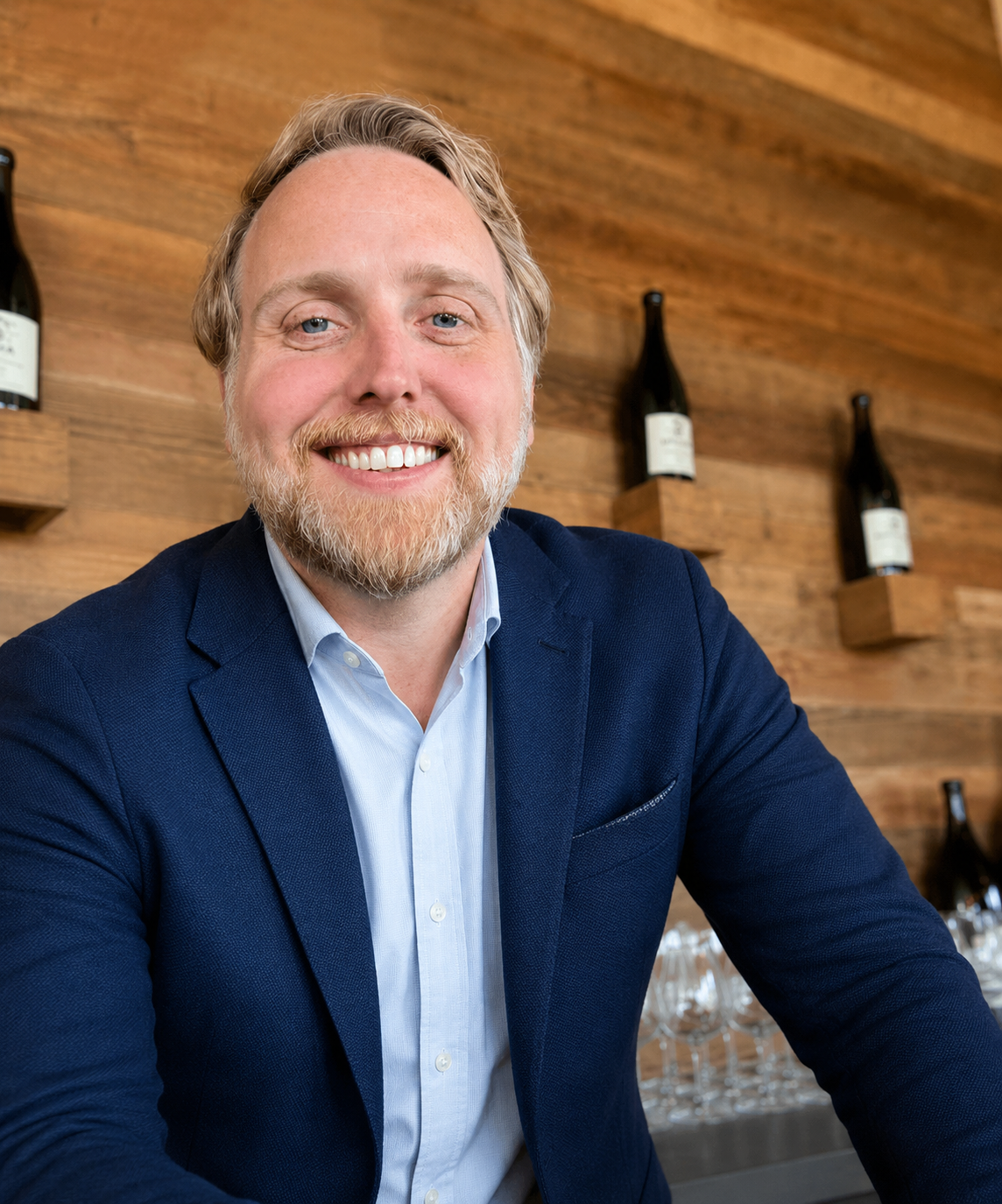 A smiling man with blonde hair and a beard, wearing a navy blue shirt, sitting in front of a wooden wall decorated with wine bottles.