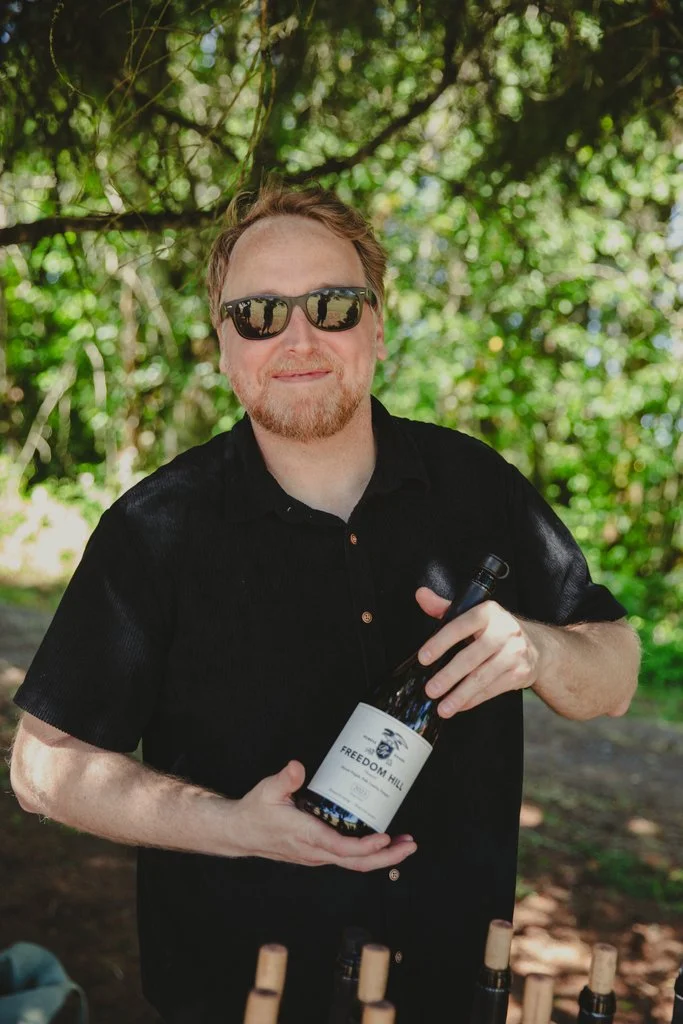 A smiling man with blonde hair and a beard, wearing a navy blue shirt, sitting in front of a wooden wall decorated with wine bottles.