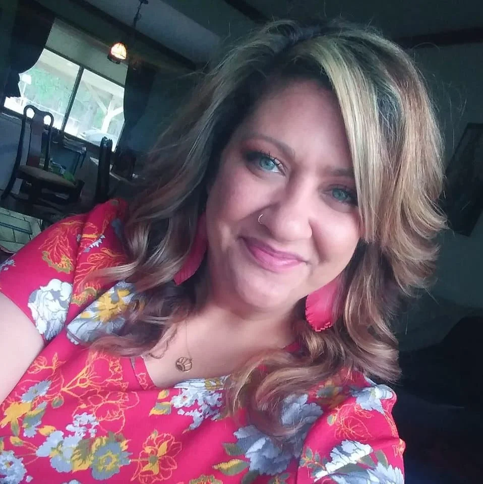 A woman with wavy, shoulder-length hair, blue eyes, wearing pink floral attire and pink earrings, smiling at the camera in a room with a window and furniture in the background.