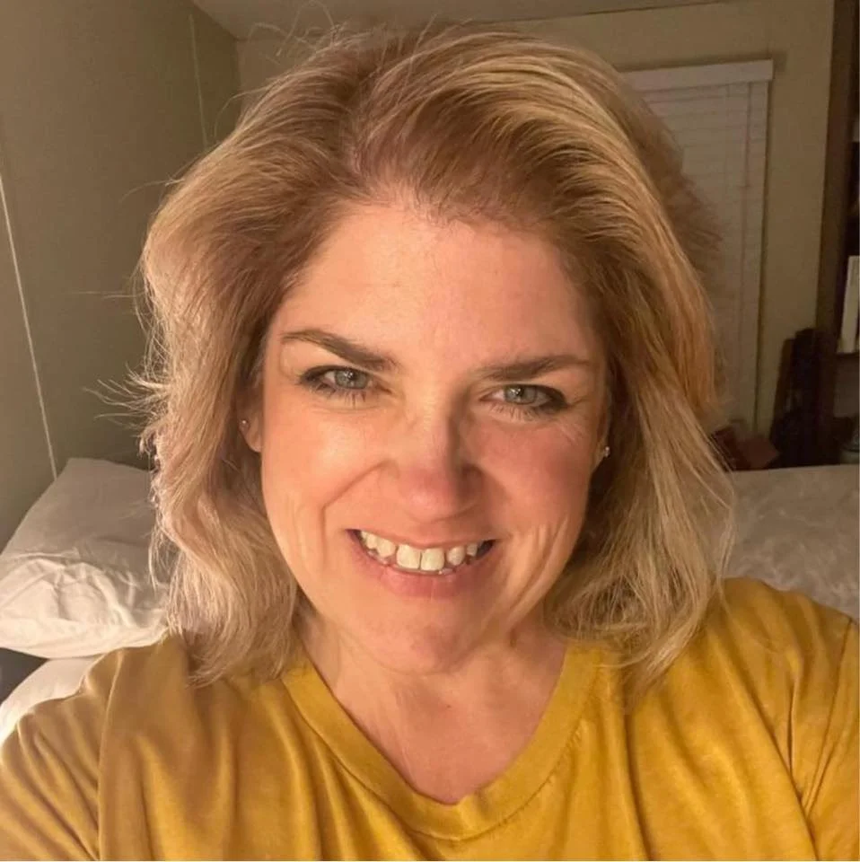 A woman with medium length blonde hair, smiling, wearing a yellow shirt, in a bedroom with a bed and window in the background.
