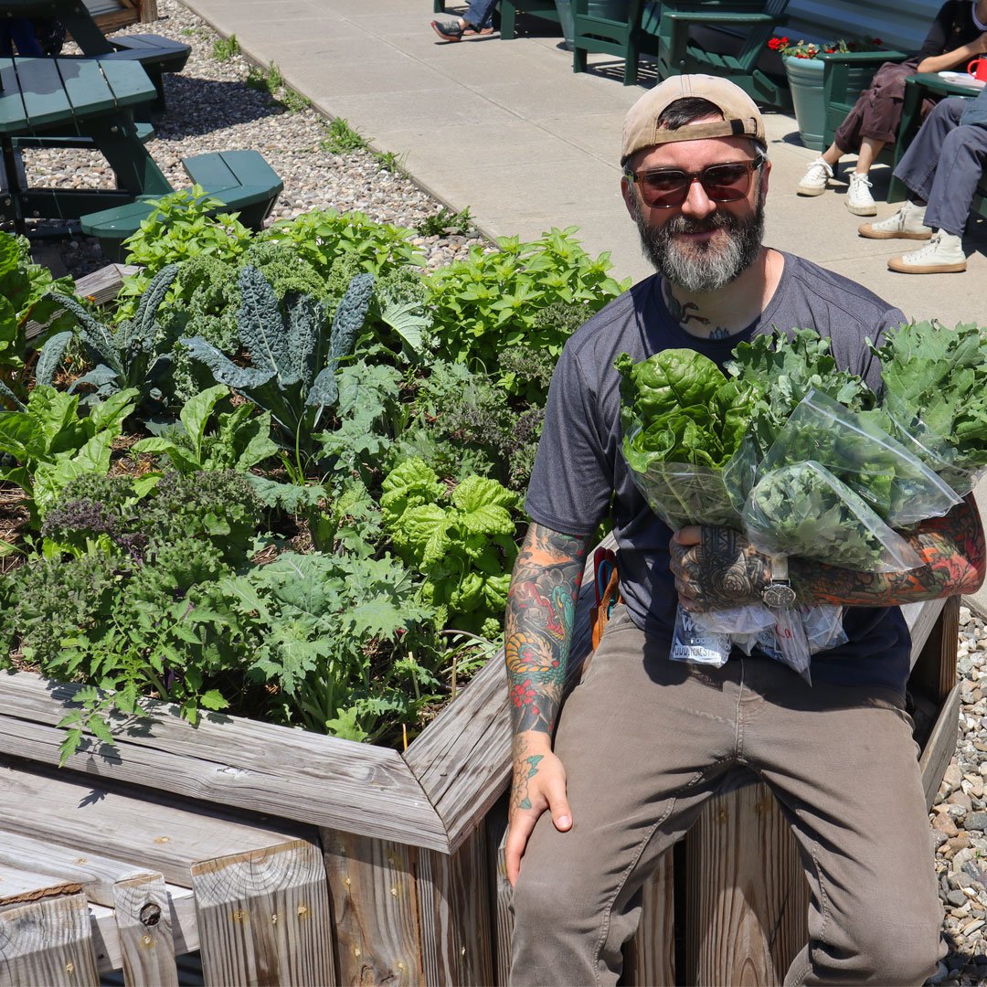 Bearded man with tattoos holding fresh greens next to garden bed