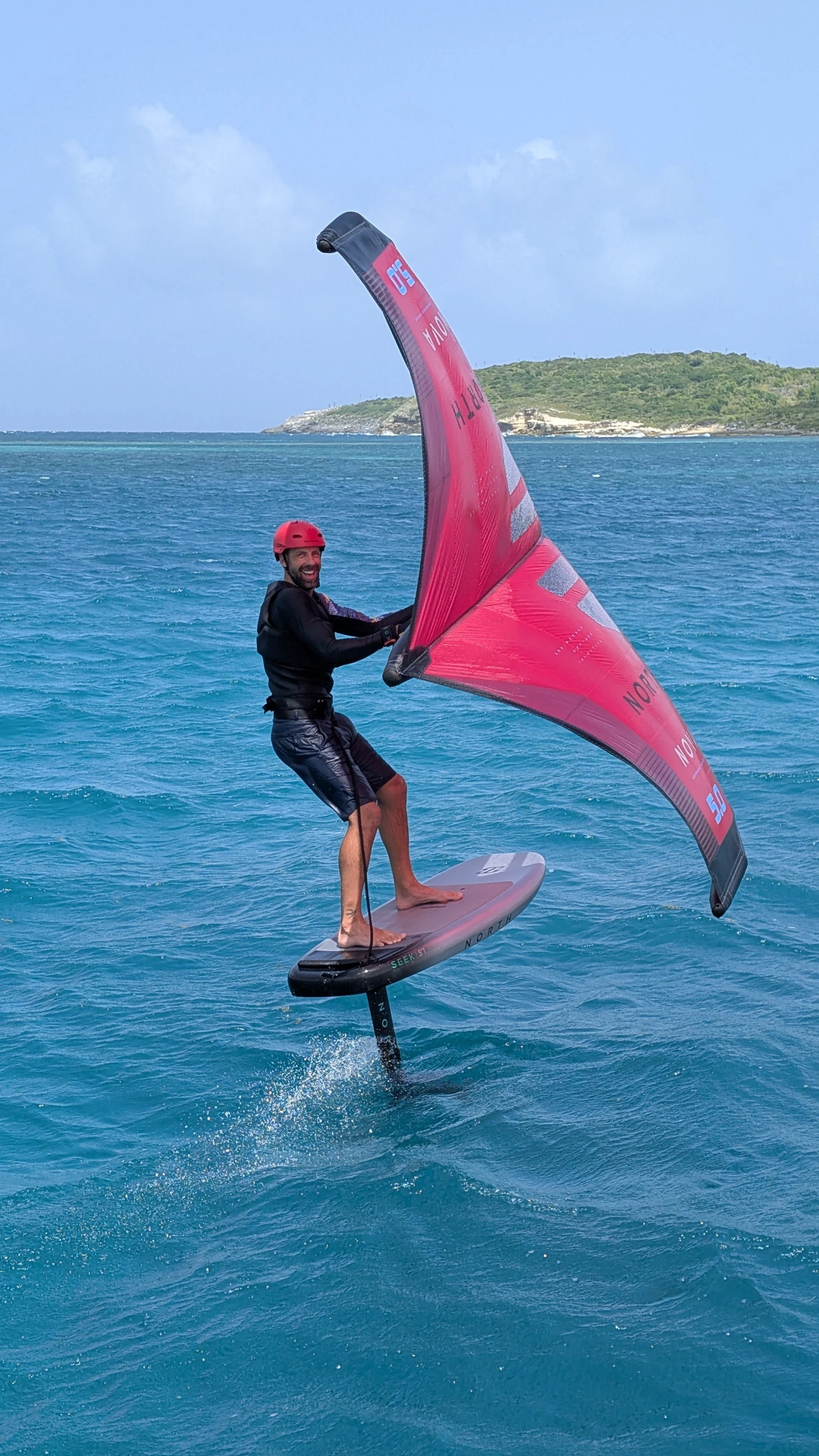 A man engaged in water sports, standing on a hydrofoil board while holding a large kiteboard in a tropical water setting with a sandy shoreline and trees in the background.