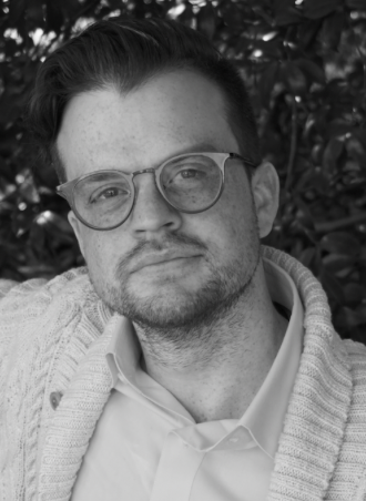 A man with glasses and facial hair poses outdoors in front of leafy foliage, wearing a light-colored collared shirt and a knitted sweater.