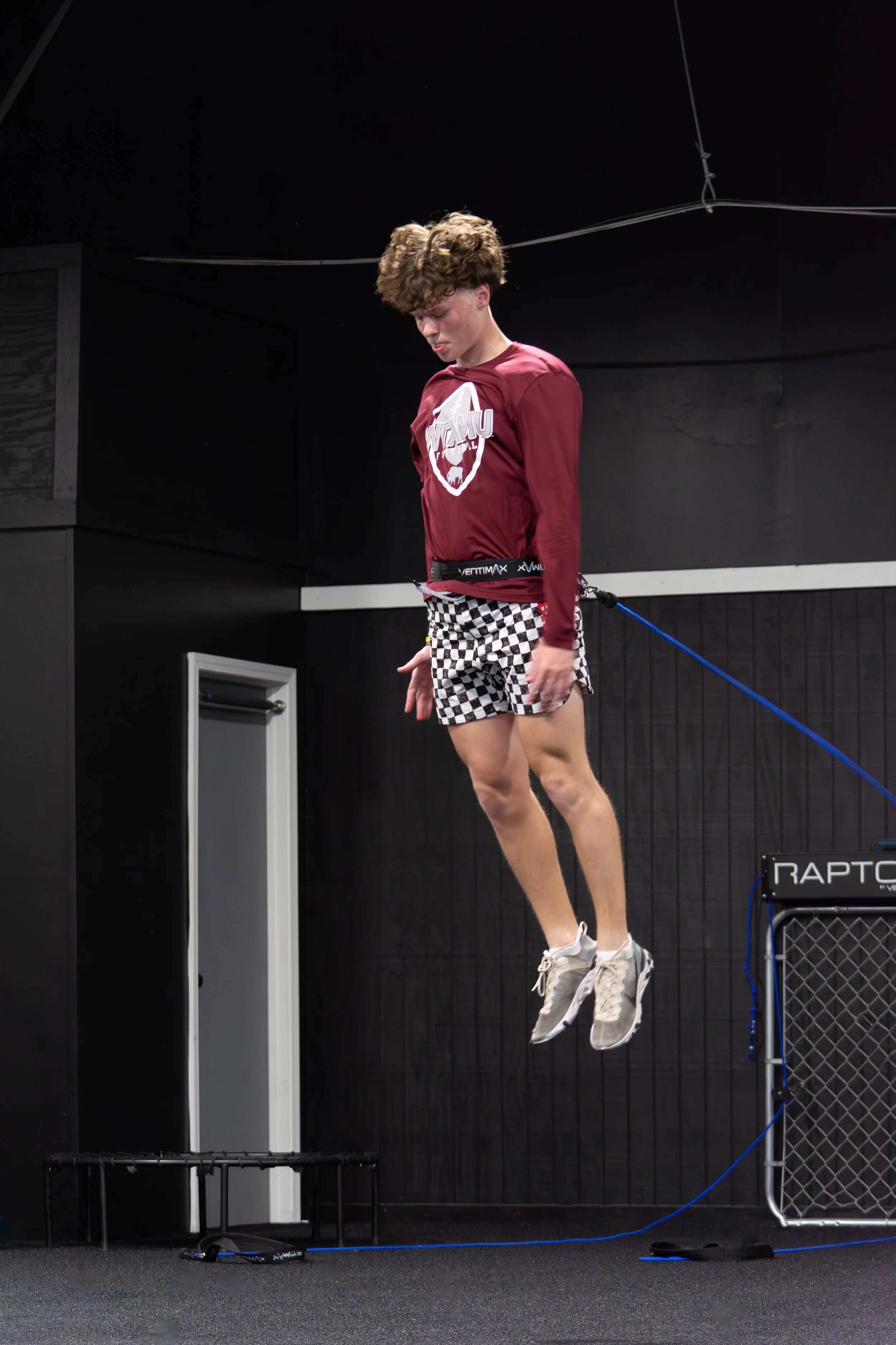 Young man in a maroon WCMU football shirt and checkered shorts jumping during an indoor athletic training exercise.