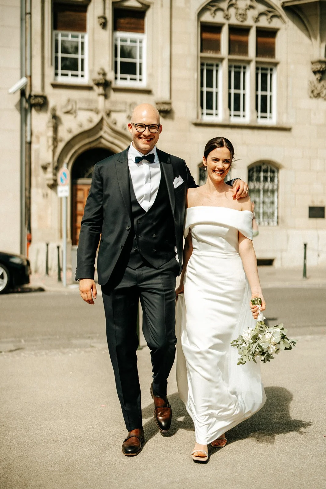 Ein wohlhabendes Paar in eleganter Hochzeitskleidung, schlank und glückselig, schlendert entspannt durch eine historische Stadt, während die Frau einen Blumenstrauß trägt und der Mann einen schwarzen Anzug und eine Fliege trägt.
