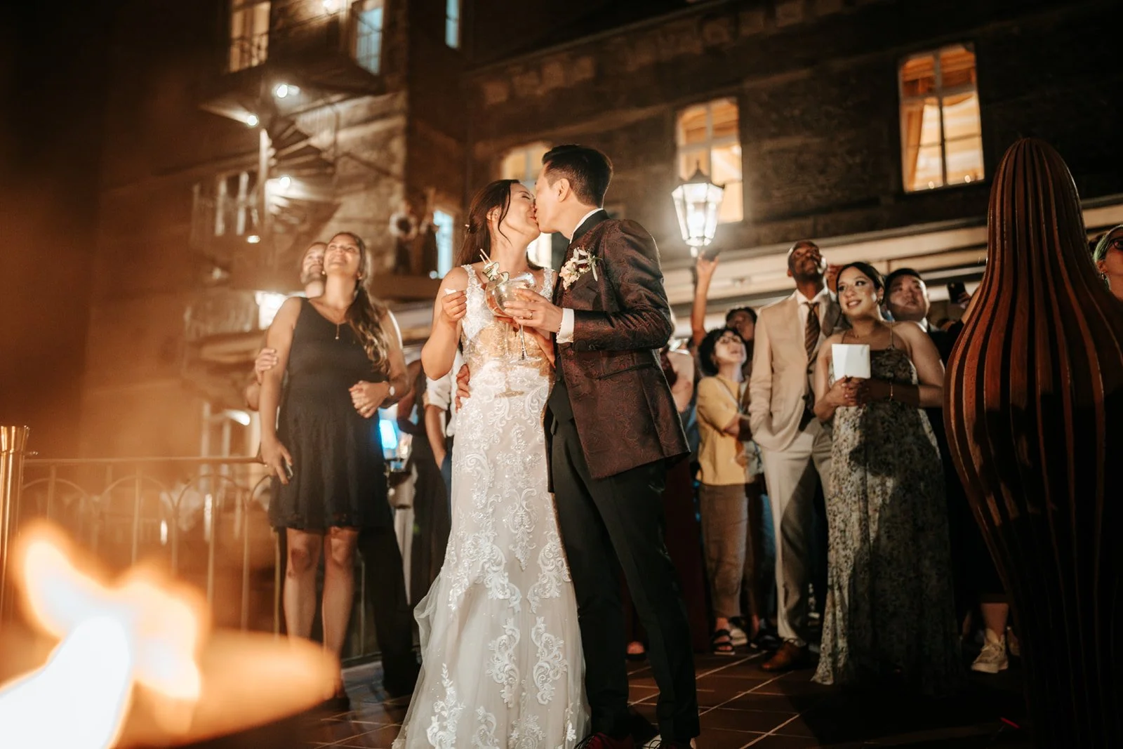 Braut und Bräutigam küssen sich während ihrer Hochzeitsfeier, umgeben von Gästen in einer festlichen Atmosphäre.