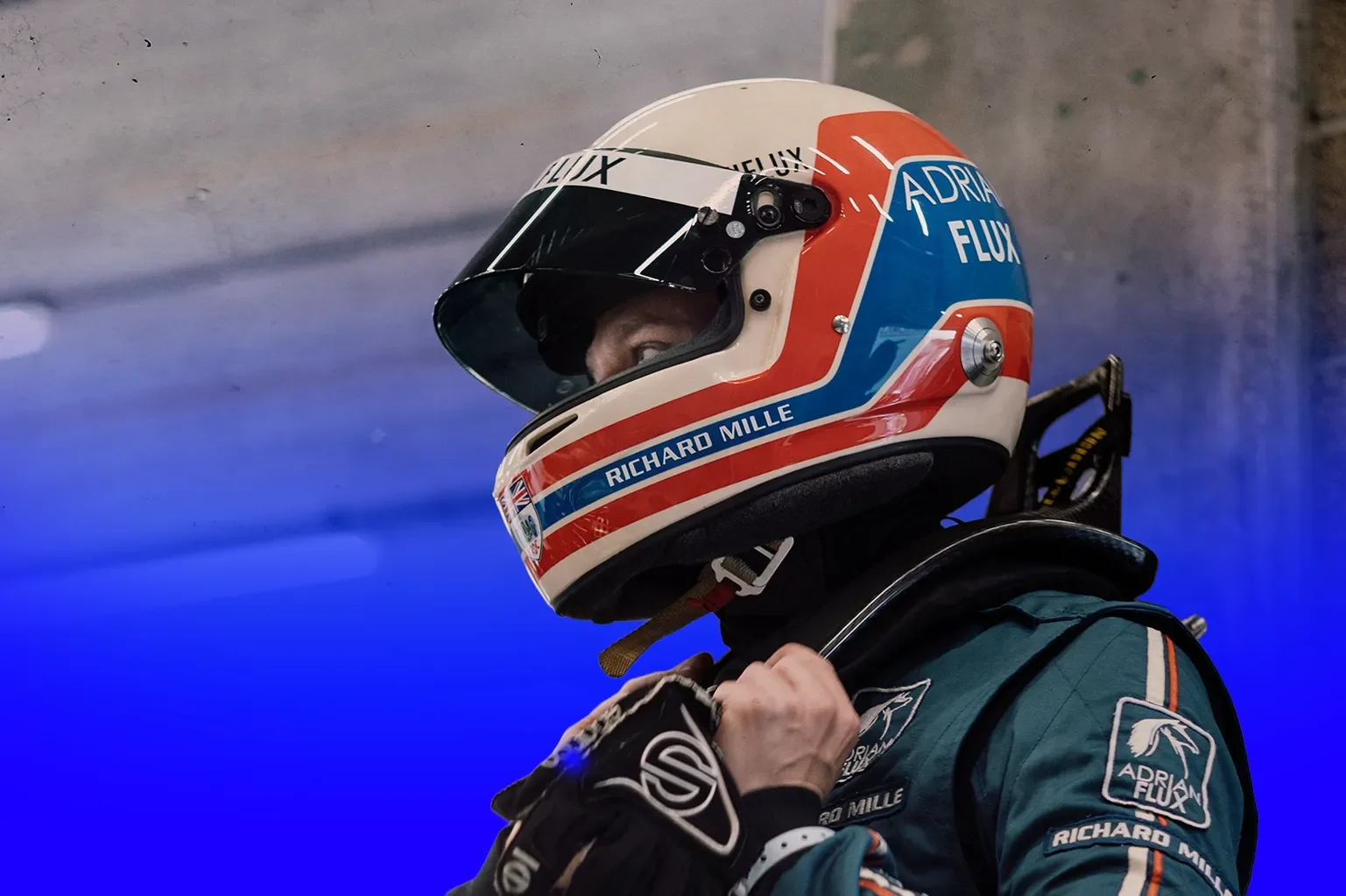 A race car driver wearing a Richard Mille helmet with an Adrien Flux logo adjusts the strap of his racing suit.