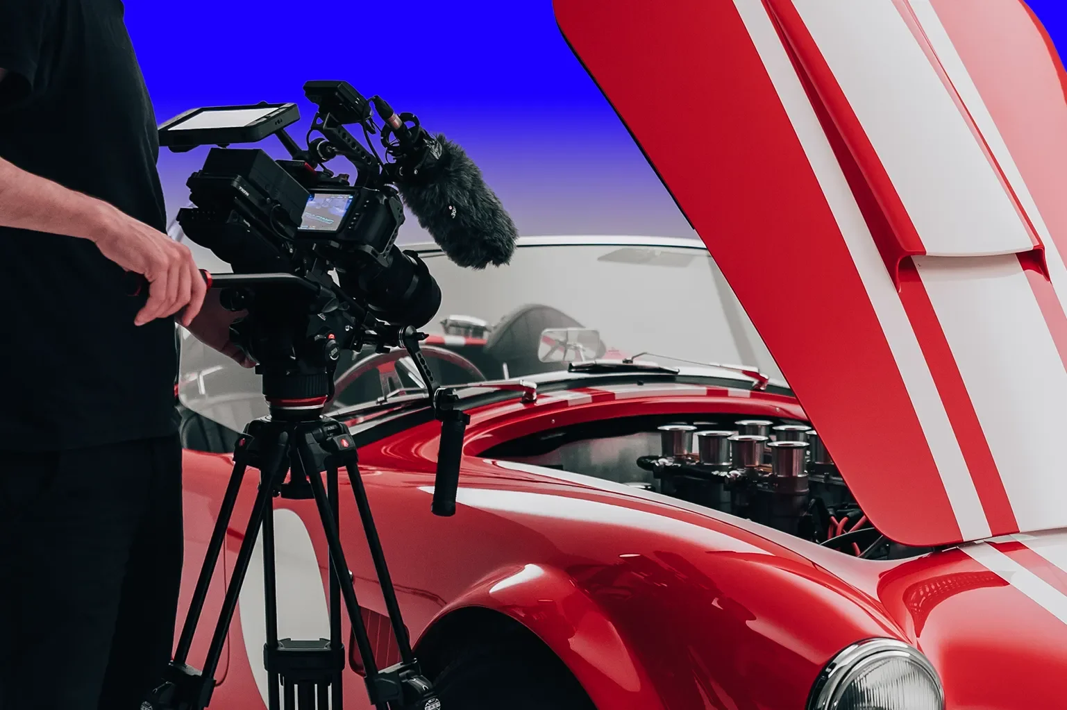 A person operating a professional video camera on a tripod next to a red vintage racing car with its hood open, revealing the engine.