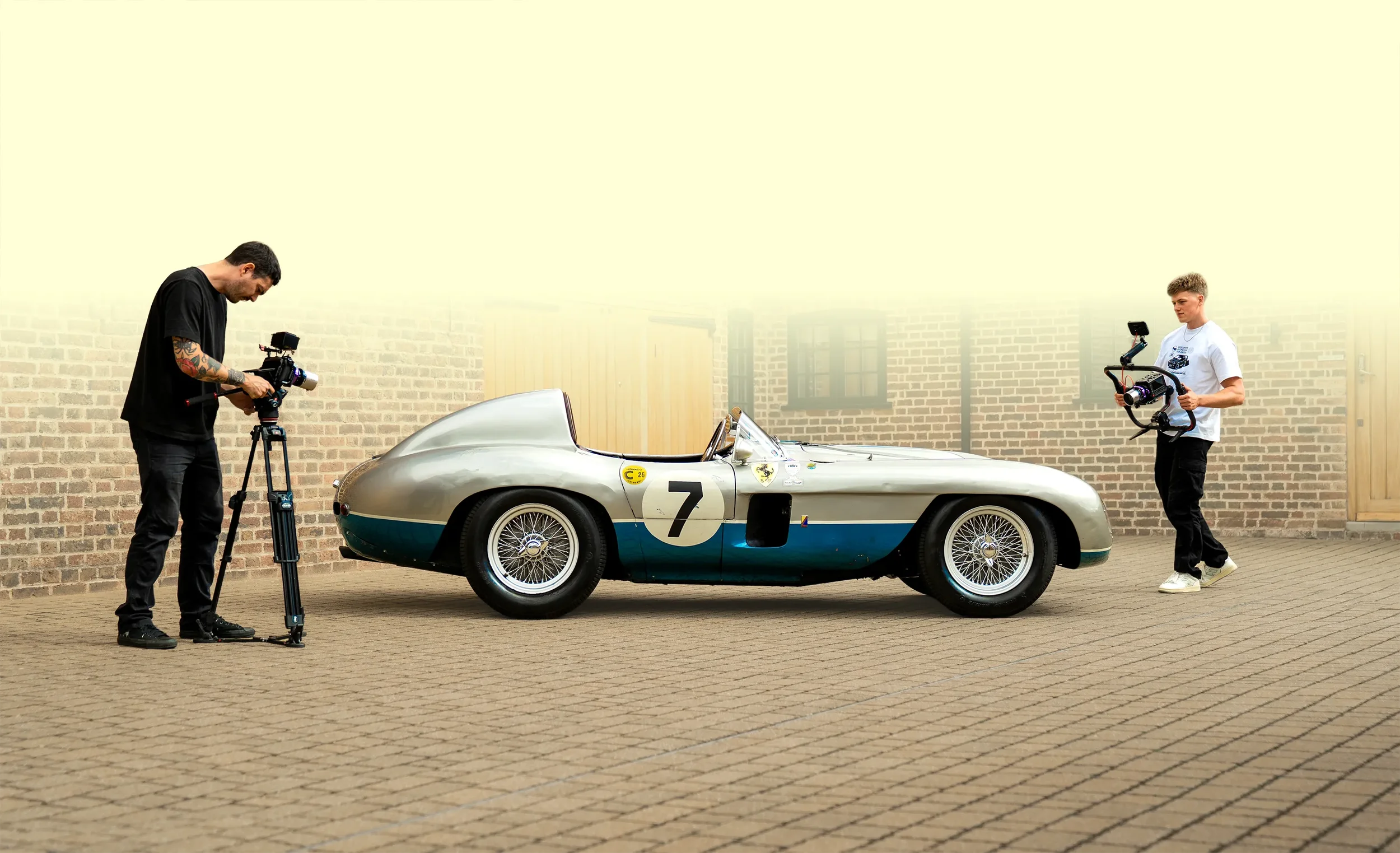 Two men filming a vintage race car with a camera mounted on a tripod. The car is silver with blue accents, marked with the number 7, and features a classic open cockpit design. They are standing outdoors against a brick wall.