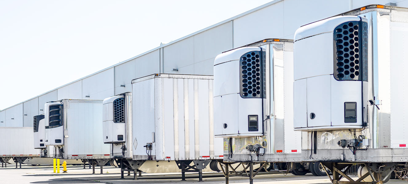 Row of white refrigerated semi-trailers parked outside of a large warehouse building.