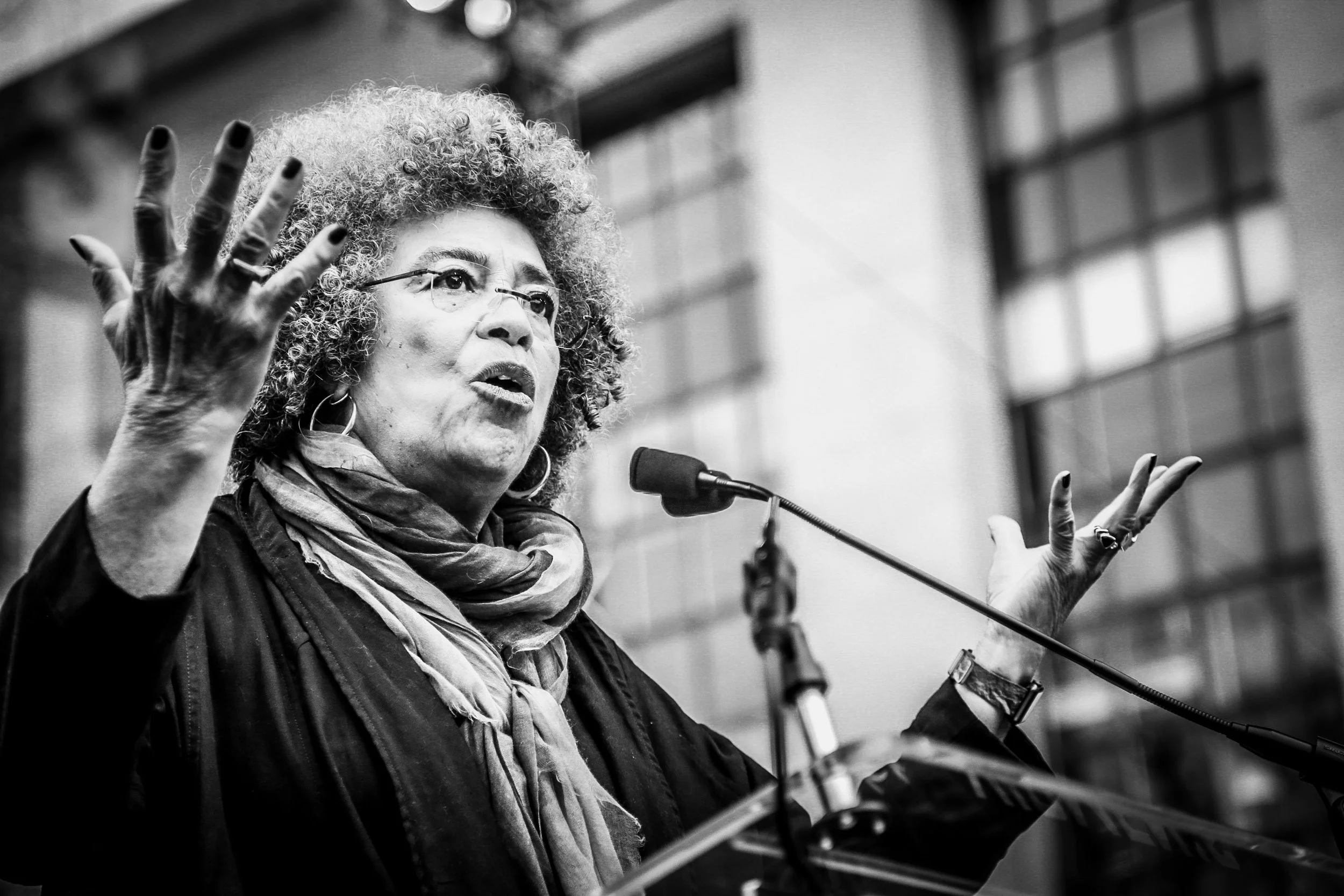 Black and white photo of a woman speaking at a podium with a microphone, gesturing with both hands, set outdoors with a building in the background.