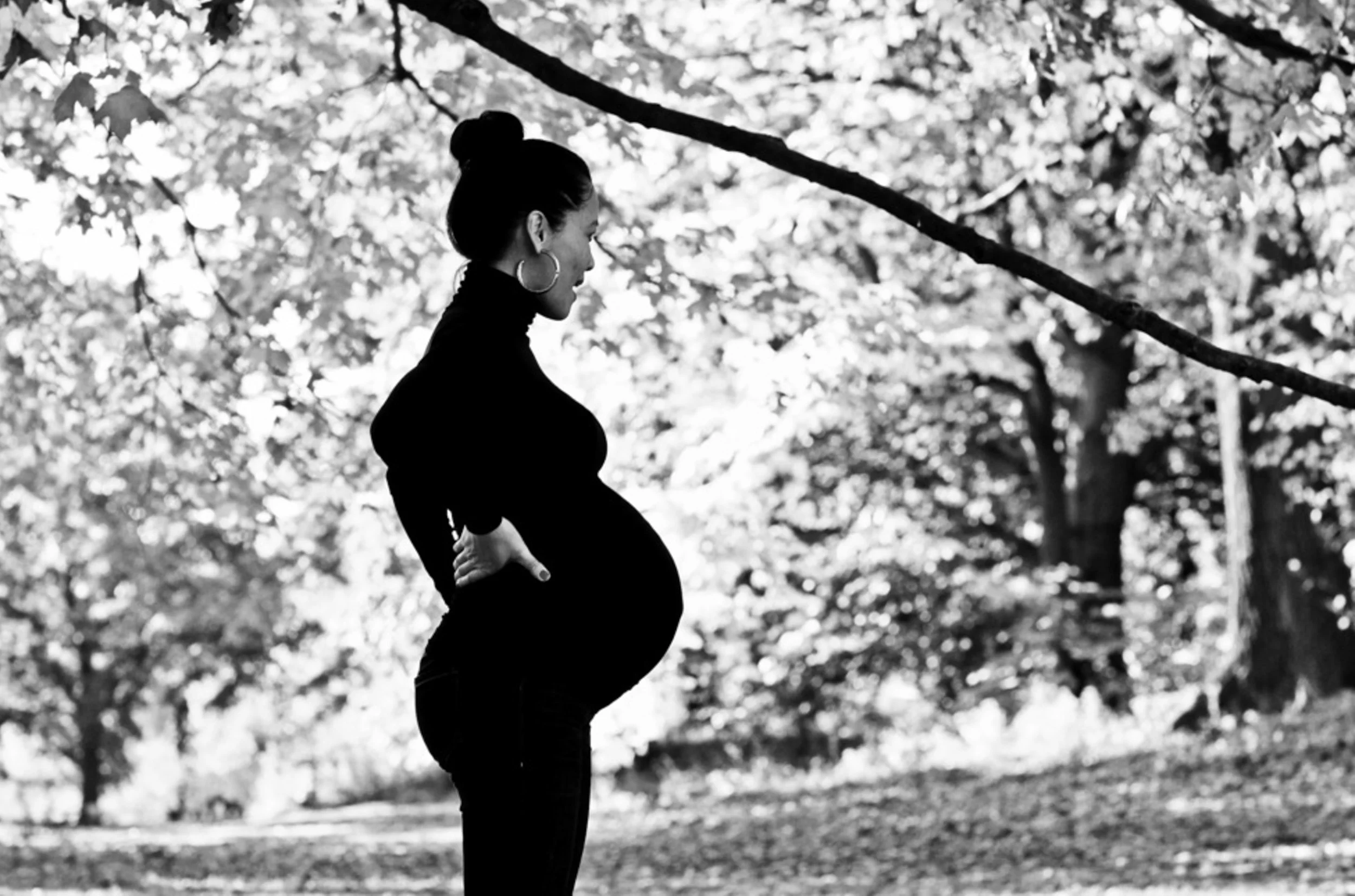 Silhouette of a pregnant woman standing outdoors in a park, looking down, with her hands on her hips, surrounded by trees in black and white.