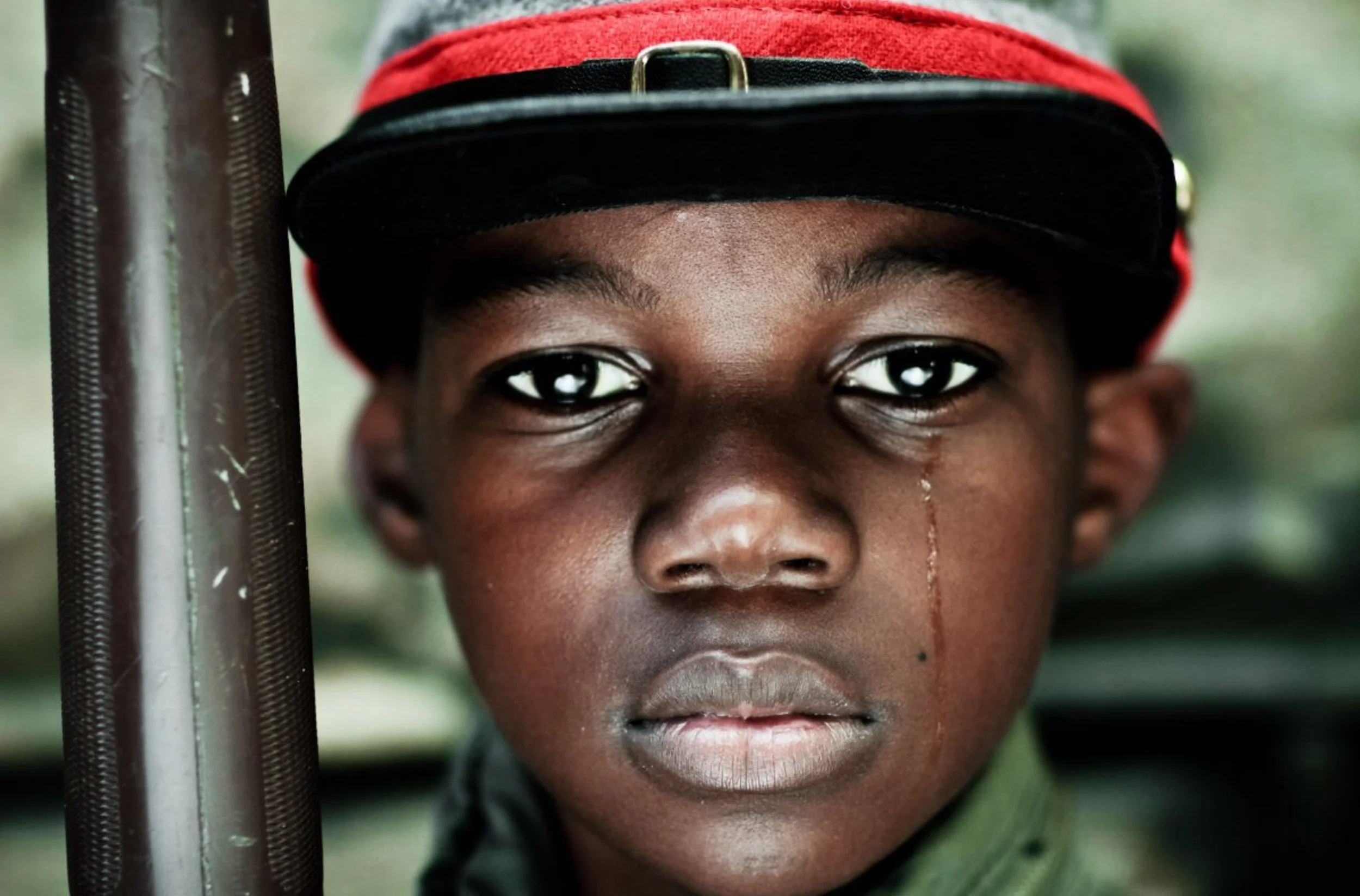 Close-up of a young boy with dark skin, wearing a red and black military cap, looking directly into the camera with intense eyes, standing near a metallic pole.