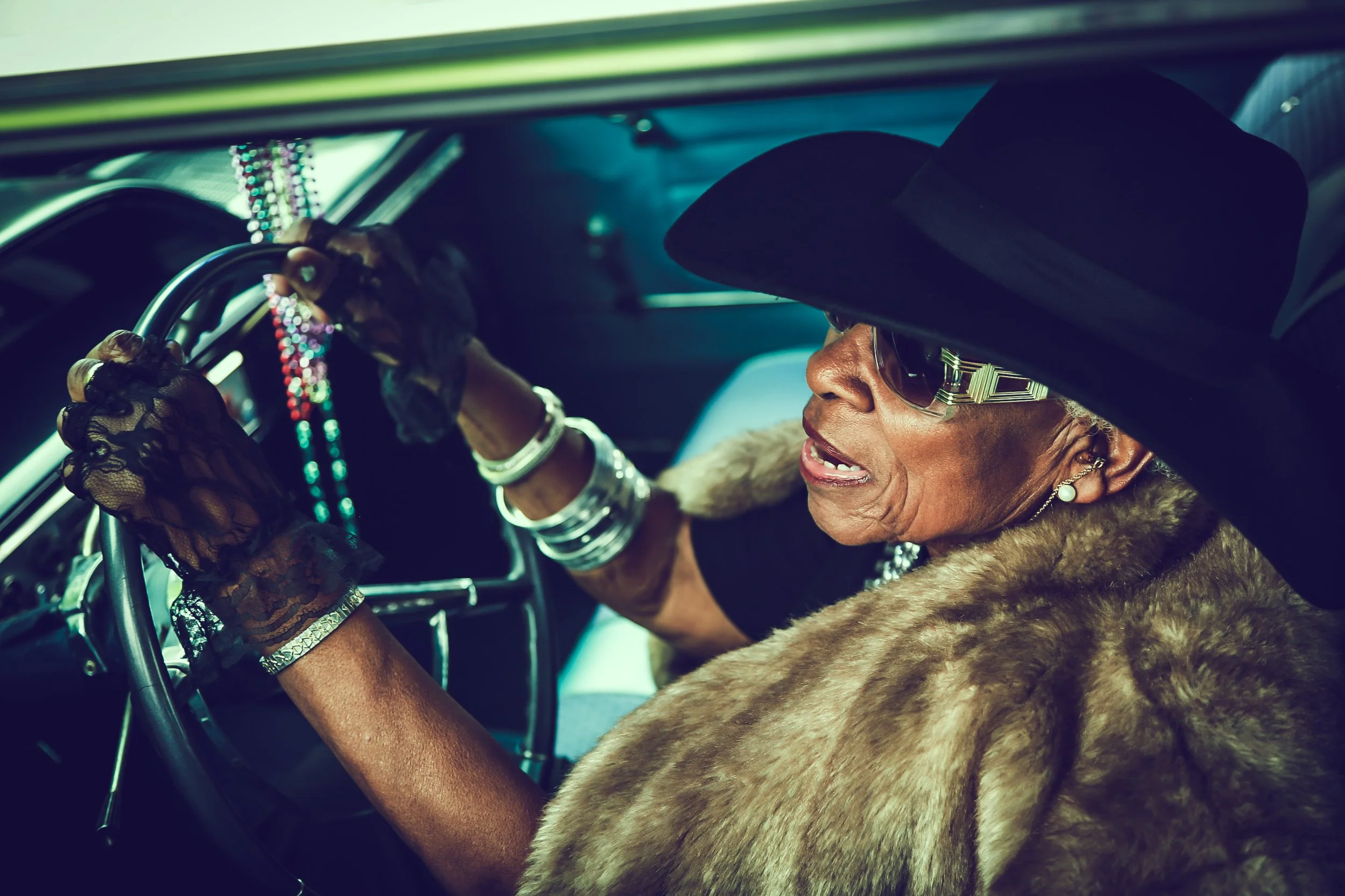 An elderly woman dressed in luxury clothing, including a fur coat, large hat, sunglasses, and jewelry, is driving a vintage car, holding the steering wheel with a determined expression.