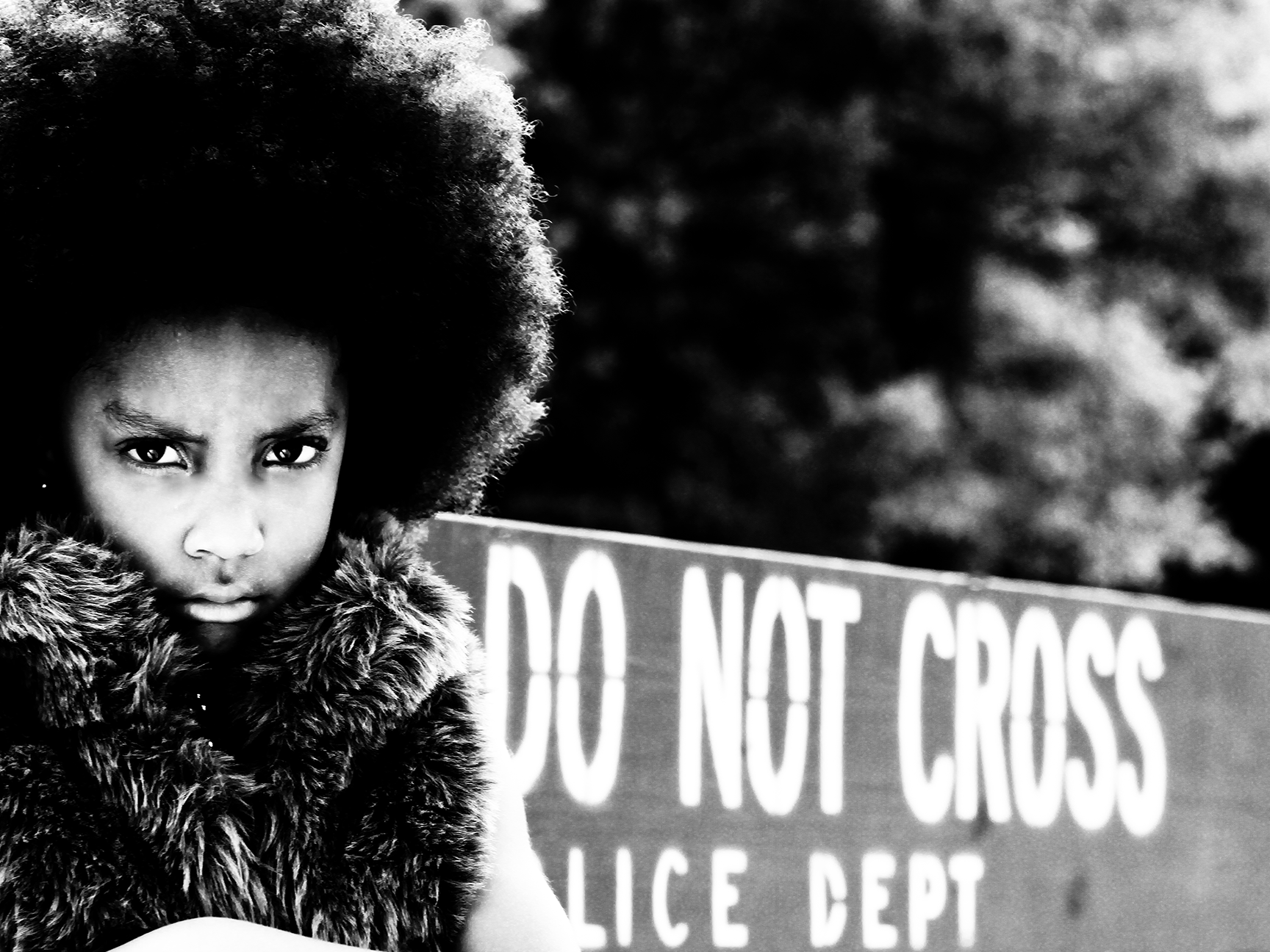 A young girl with serious expression, wearing a faux fur-lined coat, standing in front of a sign that reads 'Do Not Cross Police Dept,' with a blurred background of trees.