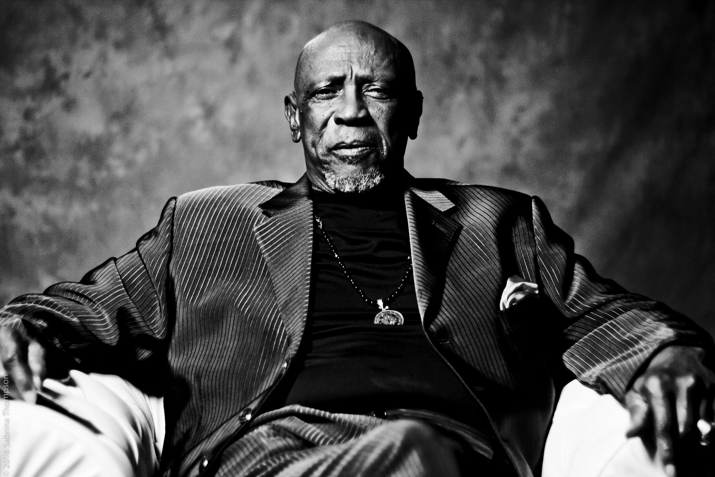 An older man with a bald head and gray beard, dressed in a pinstripe suit jacket, black shirt, and white pants, sitting in a chair against a textured background.