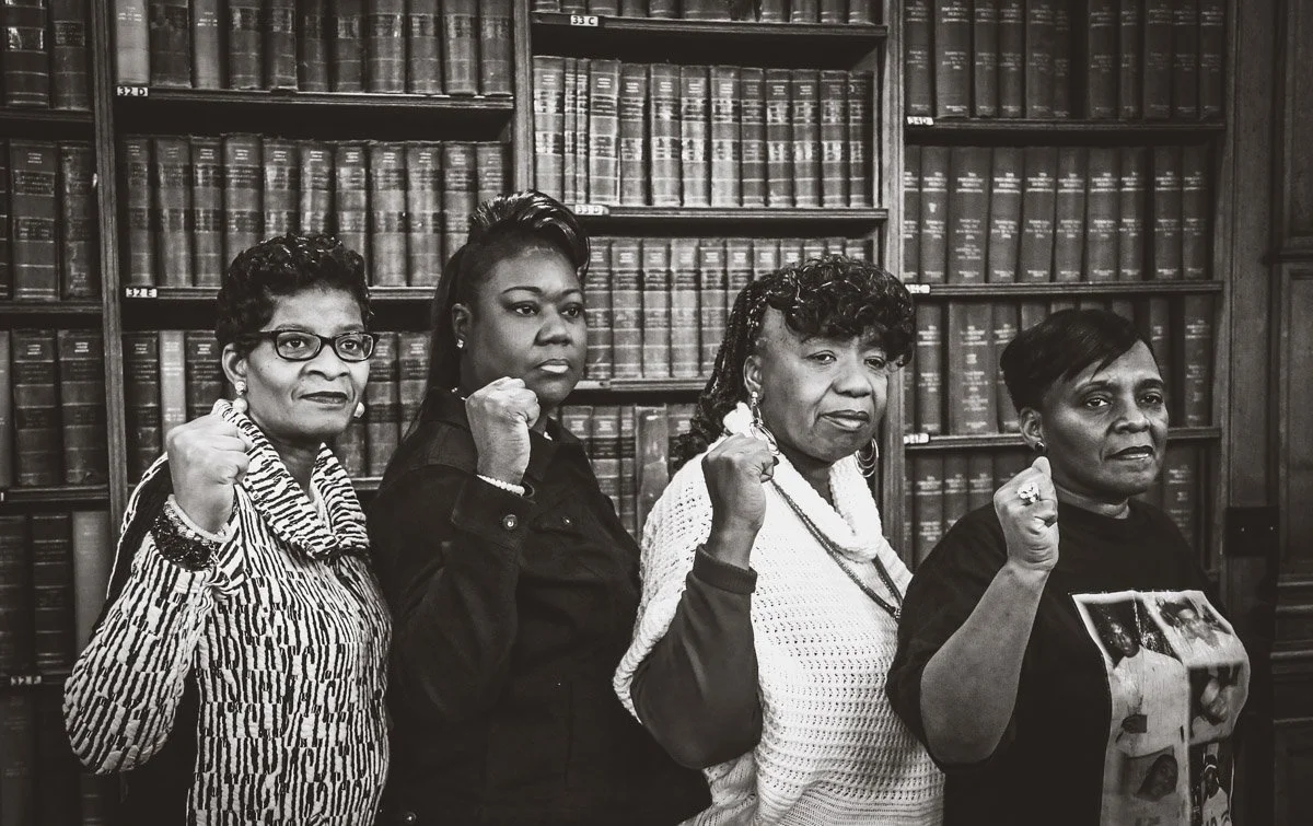 Four women standing in a library, each with one arm raised and clenched fist, posing with serious expressions.