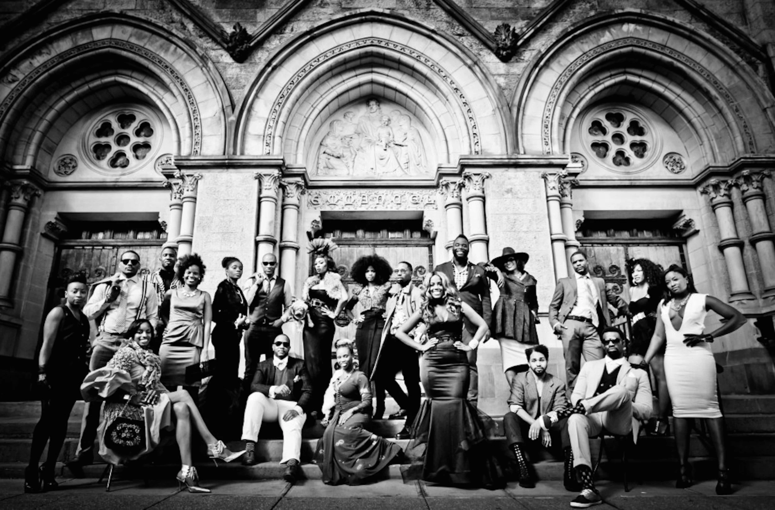 A black and white photo of a diverse group of people posing on the steps of a historic building with detailed stone architecture and arched windows.