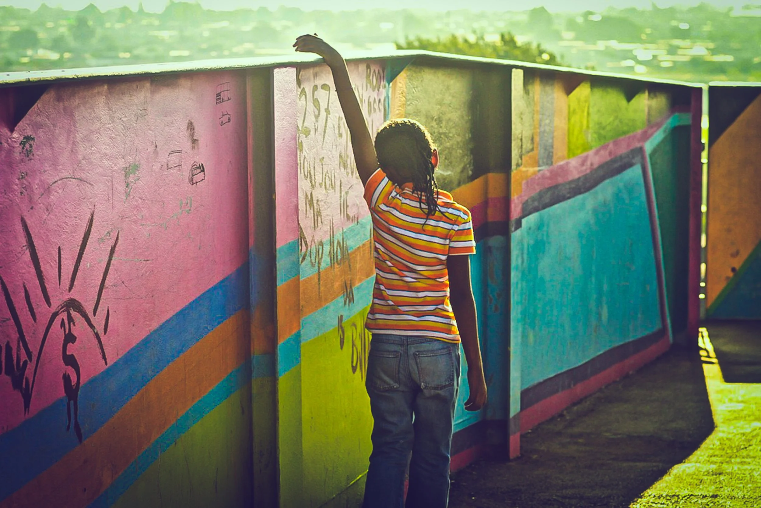 A girl in a striped shirt and jeans is painting or drawing on a colorful mural-covered wall during sunset.