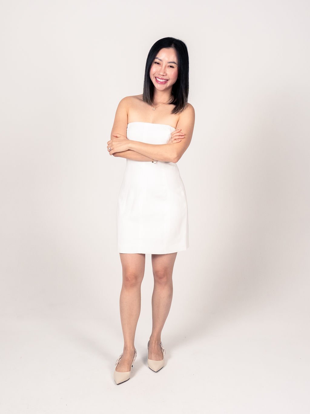 A woman with black hair in a white strapless dress, smiling with arms crossed, standing against a plain white background.