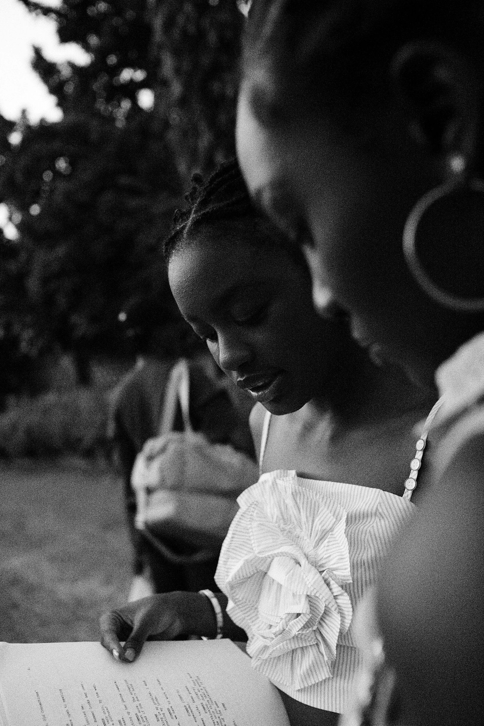 A close-up black and white photo of a woman with braided hair and earrings, wearing a striped shirt with ruffles on the sleeves, reading a document while outdoors, with a blurred background of trees.