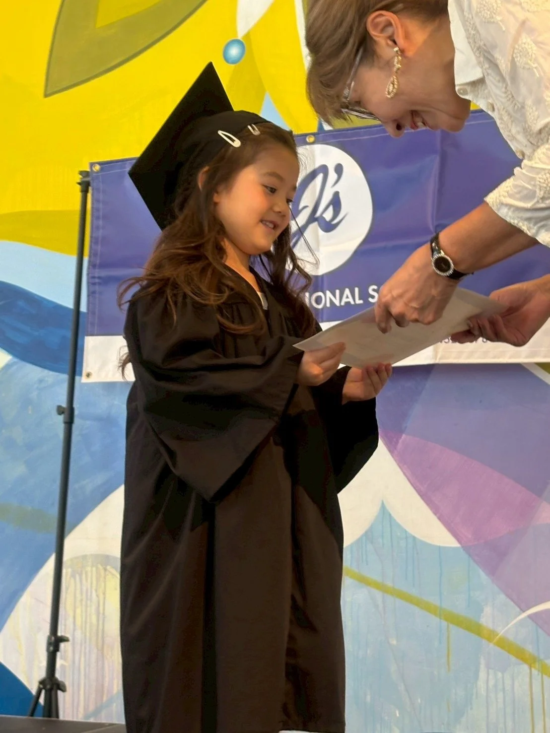 Young girl in a graduation cap and gown receiving a diploma from an older woman during a graduation ceremony.