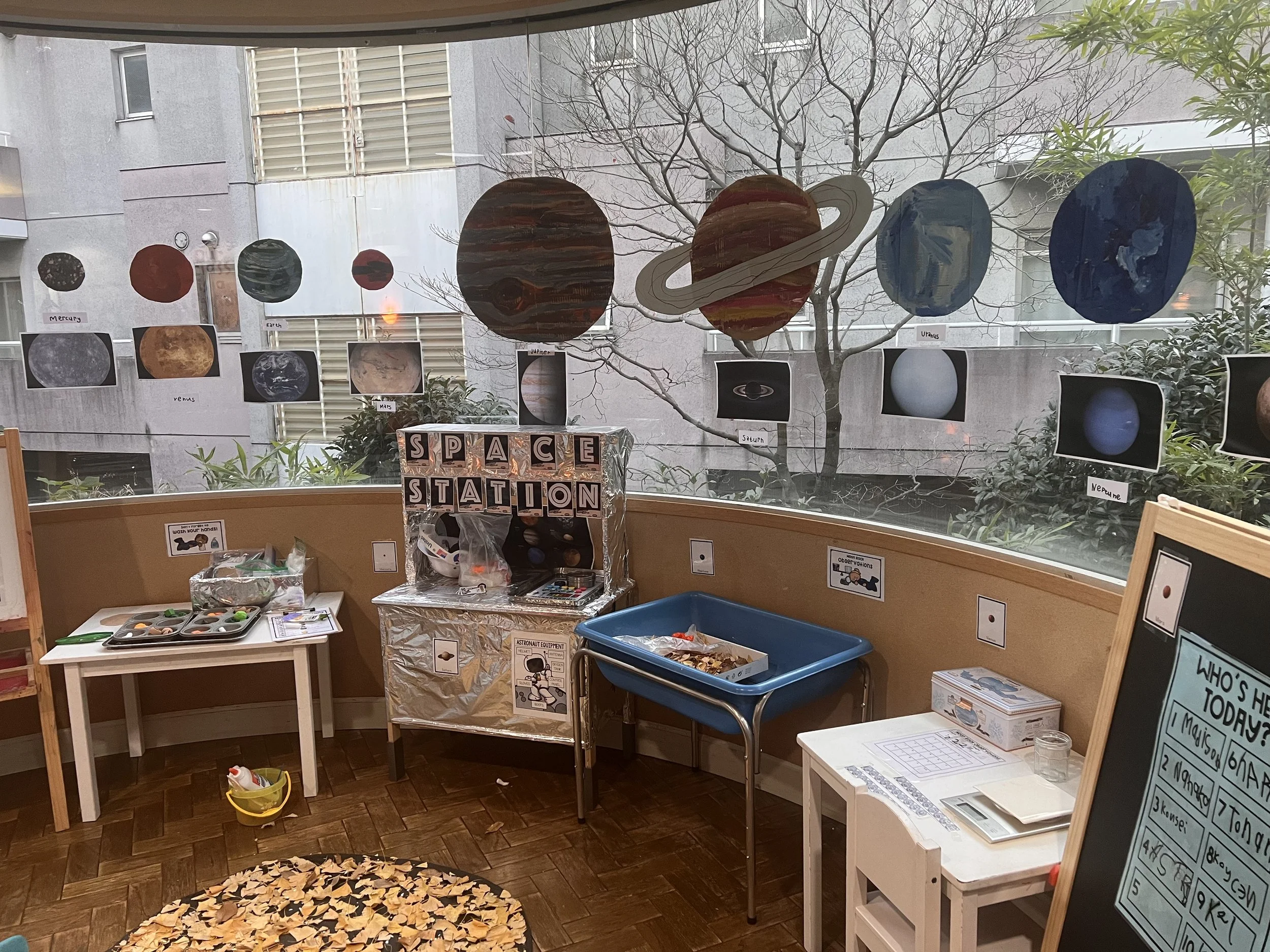 Educational display about the solar system and planets, with cutouts of planets like Mercury, Venus, Earth, Mars, Jupiter, Saturn, Uranus, and Neptune hanging in front of a window. There are tables with educational materials and a sign labeled 'Space Station'.