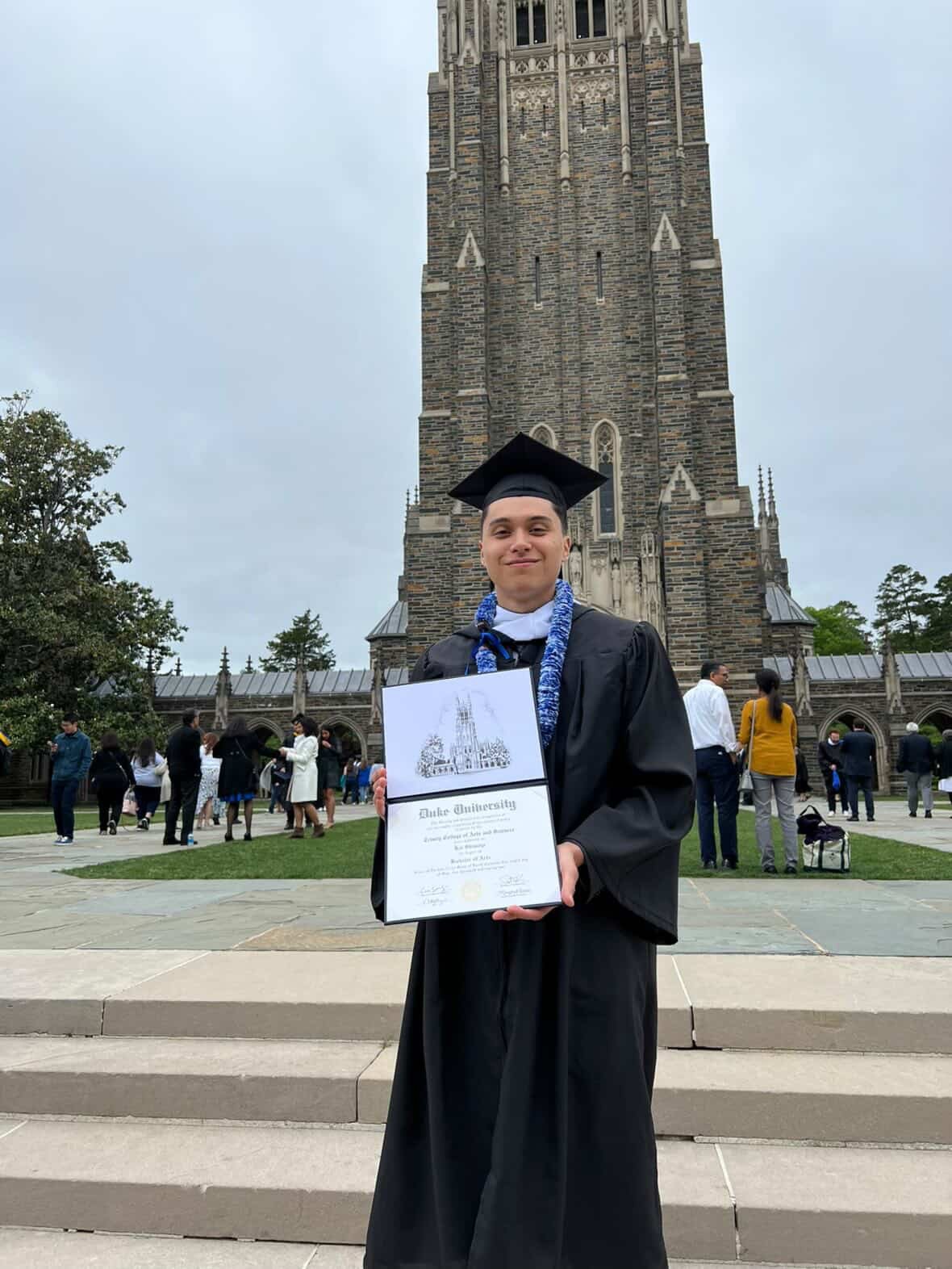 A young man in a graduation cap and gown holding a diploma in front of Duke University's iconic chapel, with other graduates and attendees gathered on the steps behind him during the day.