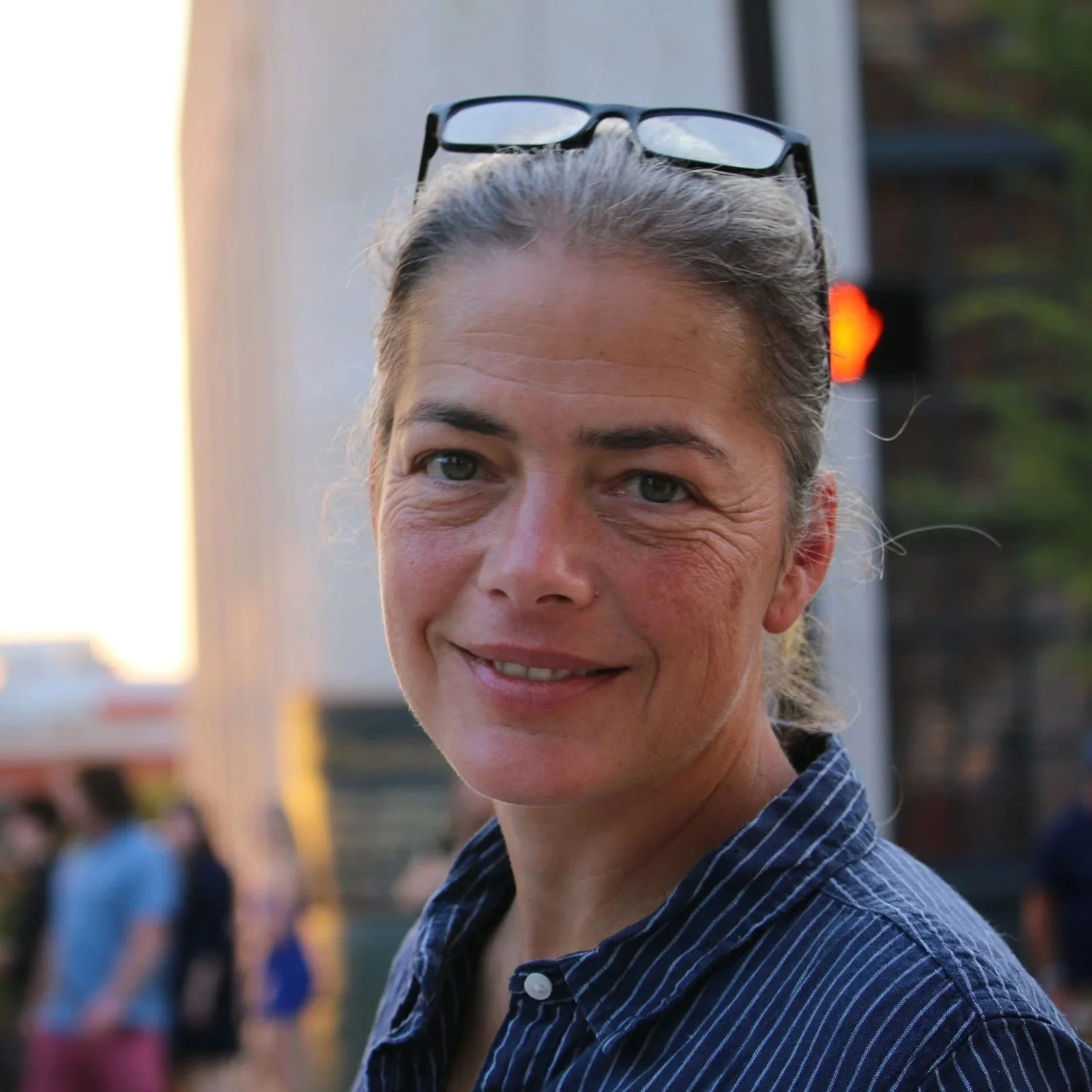 A woman with gray hair tied back, wearing glasses on her head, smiling outdoors during sunset, with a cityscape and traffic light in the background.