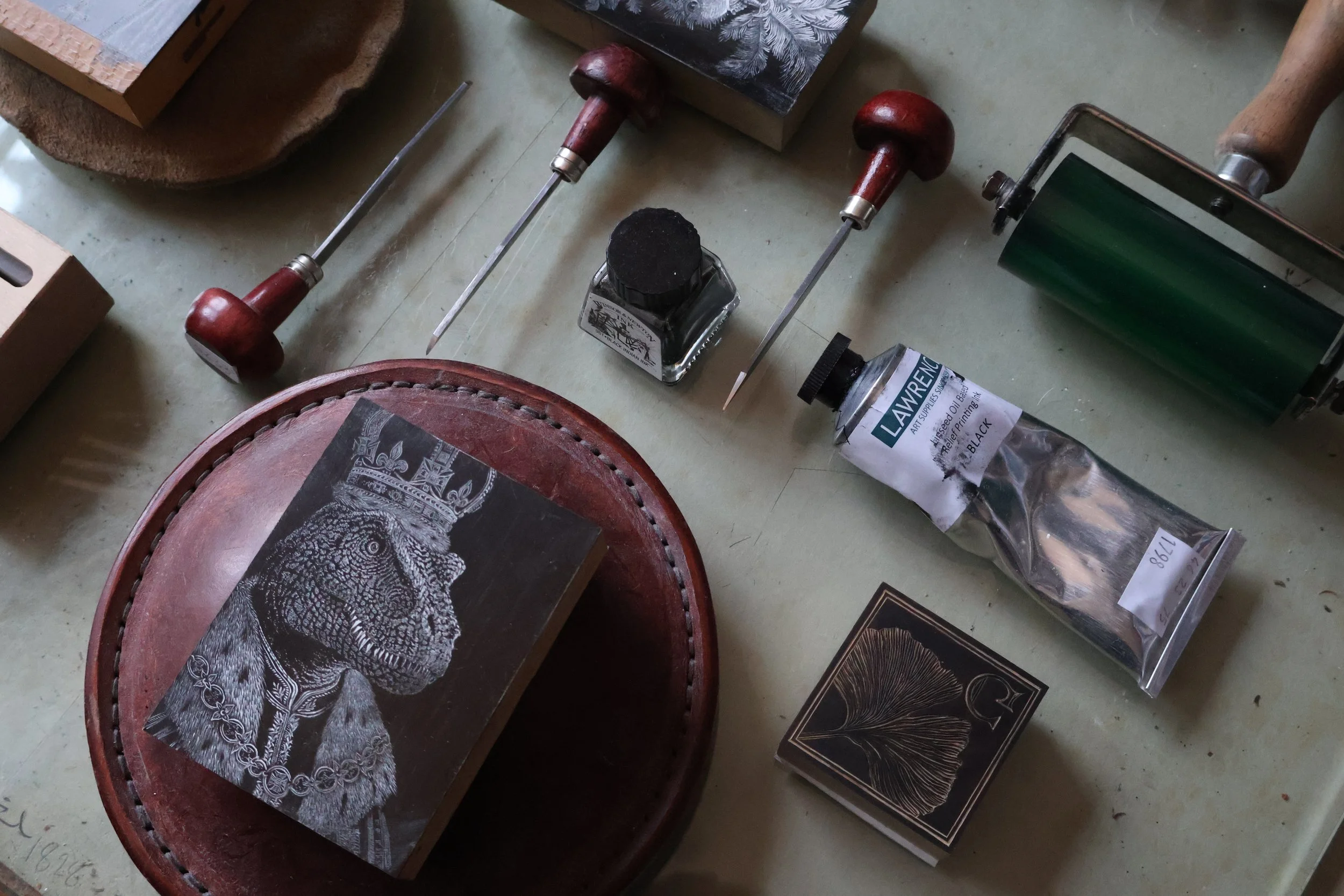 A work table with various printing tools and supplies, including a carved block with a lion wearing a crown, a roller, a bottle of ink labeled 'Lawrence', and other printing-related equipment.