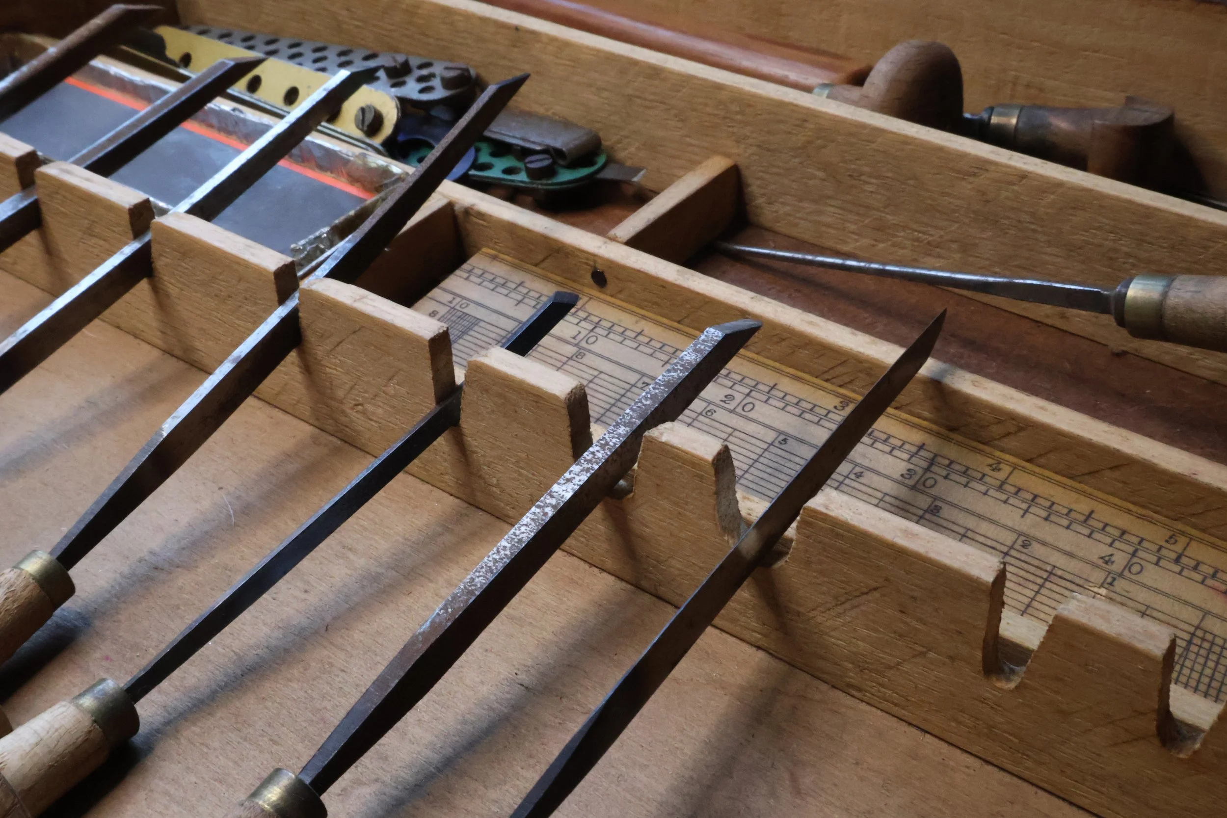 Set of woodworking chisels with wooden handles resting on a wooden tool rack with slots, inside a woodworking shop.